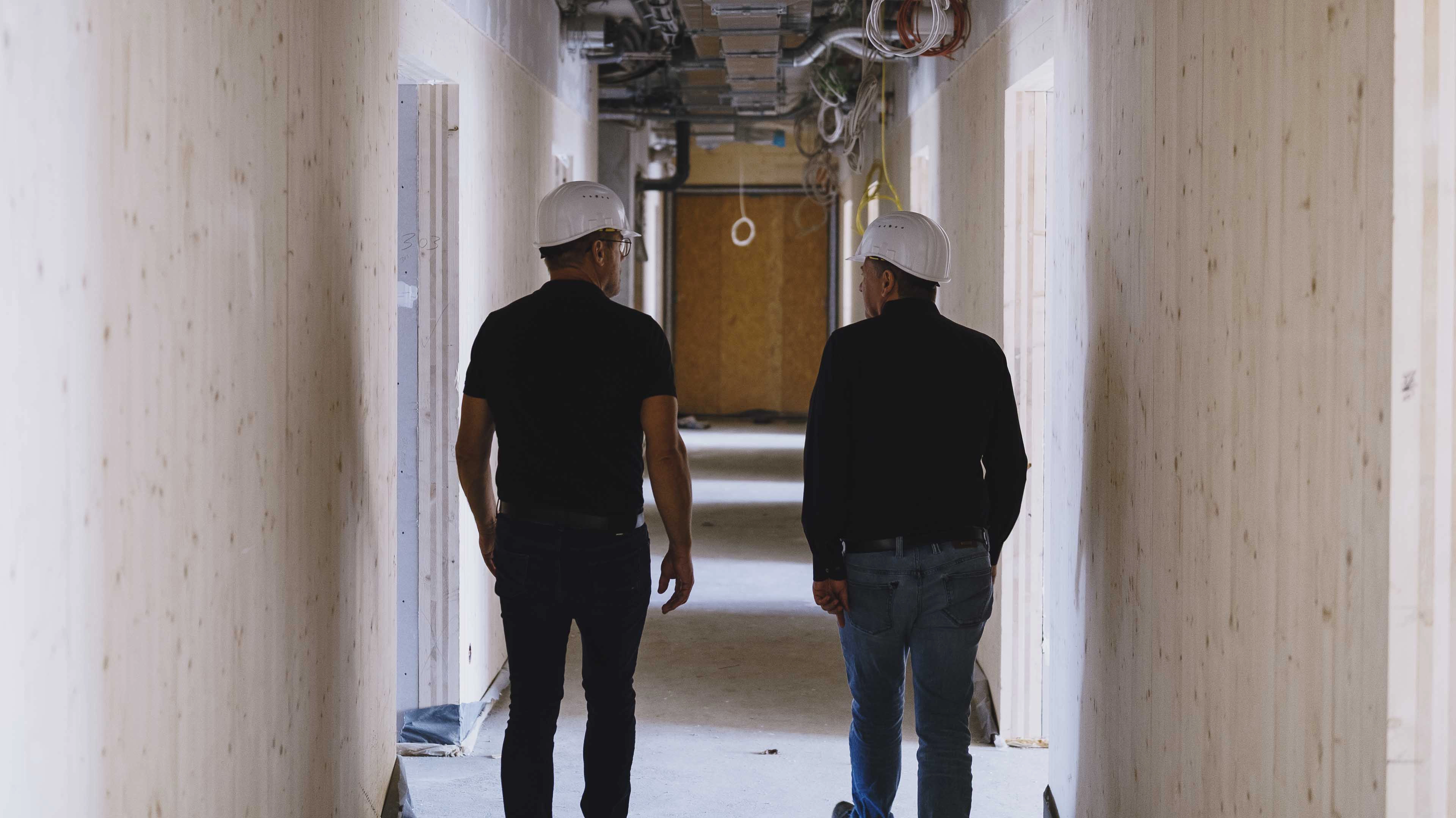 Two men wearing white construction helmets walking down a hallway under construction with exposed wiring on the ceiling.