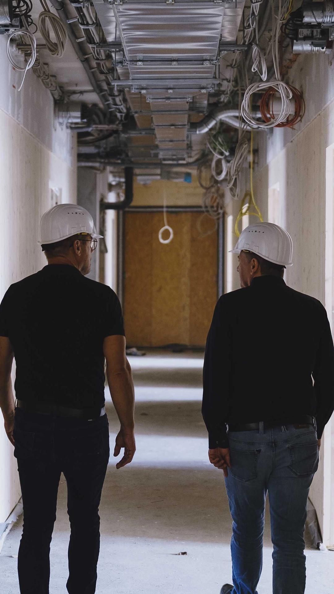 Two construction workers wearing white hard hats walking through a building corridor under renovation with visible wiring and ductwork overhead.