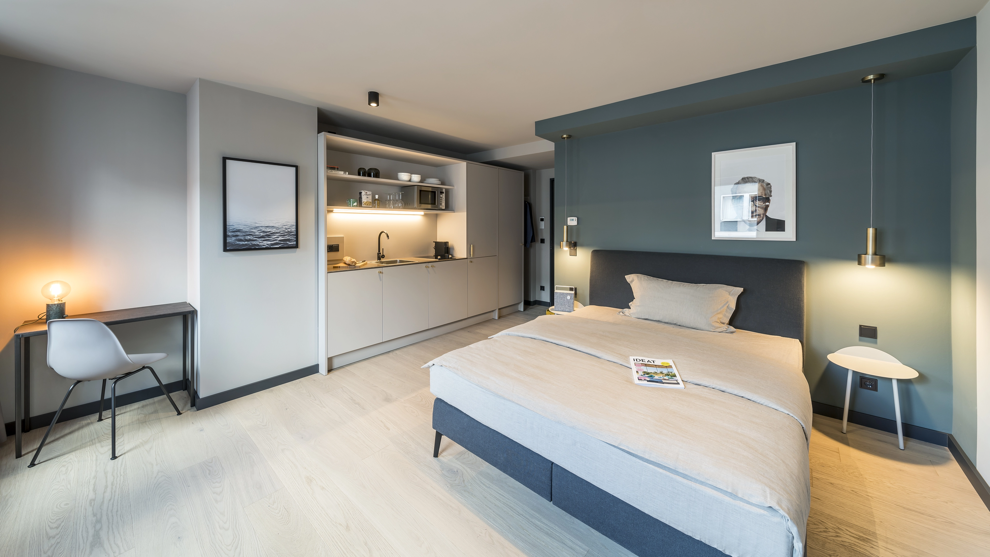 Modern hotel room with a double bed, beige bedding, minimalist kitchenette, desk with lamp, and framed artworks on blue and white walls.