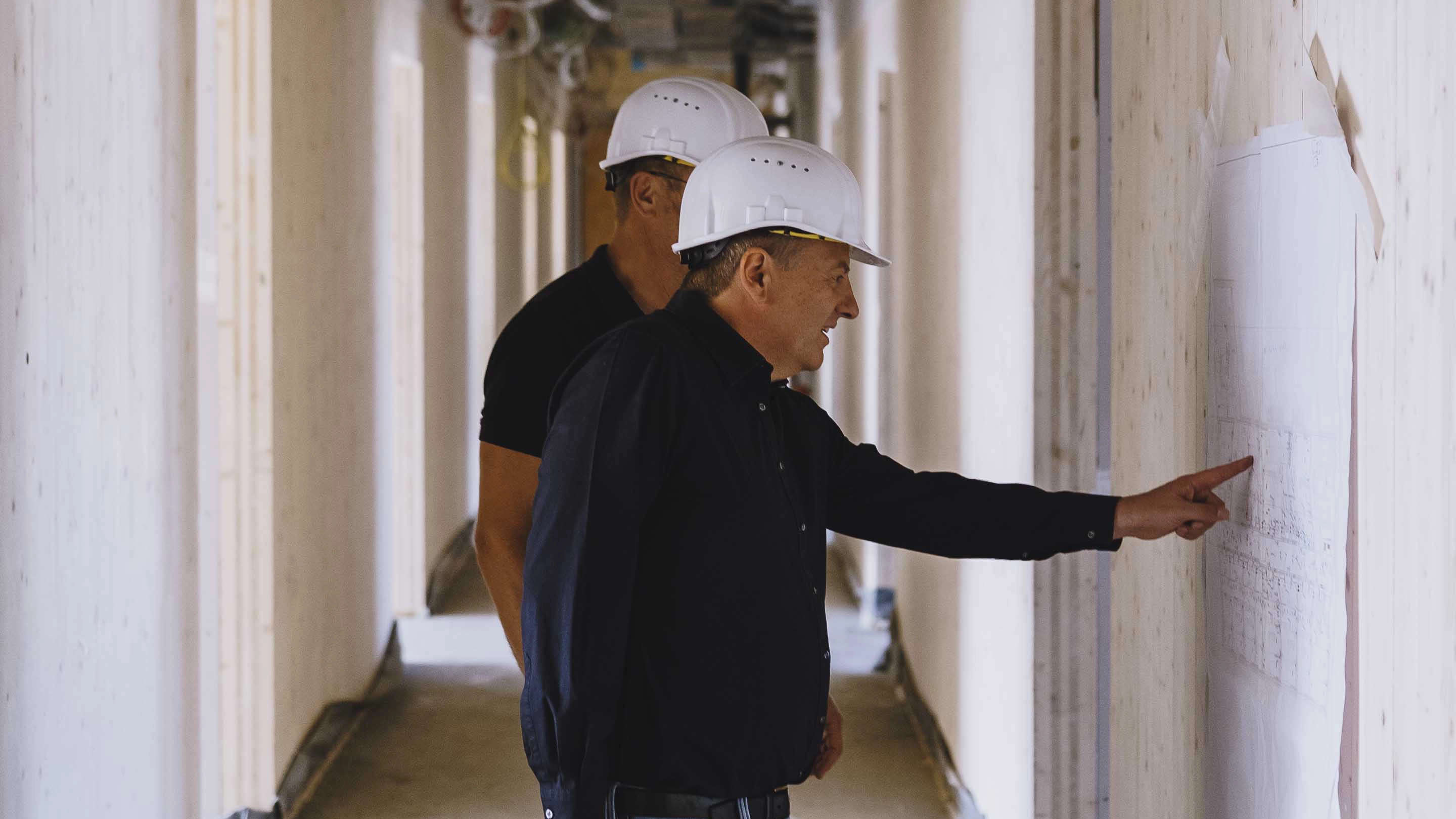Two construction workers wearing white hard hats examining and pointing at a blueprint on a wall inside a building under construction.