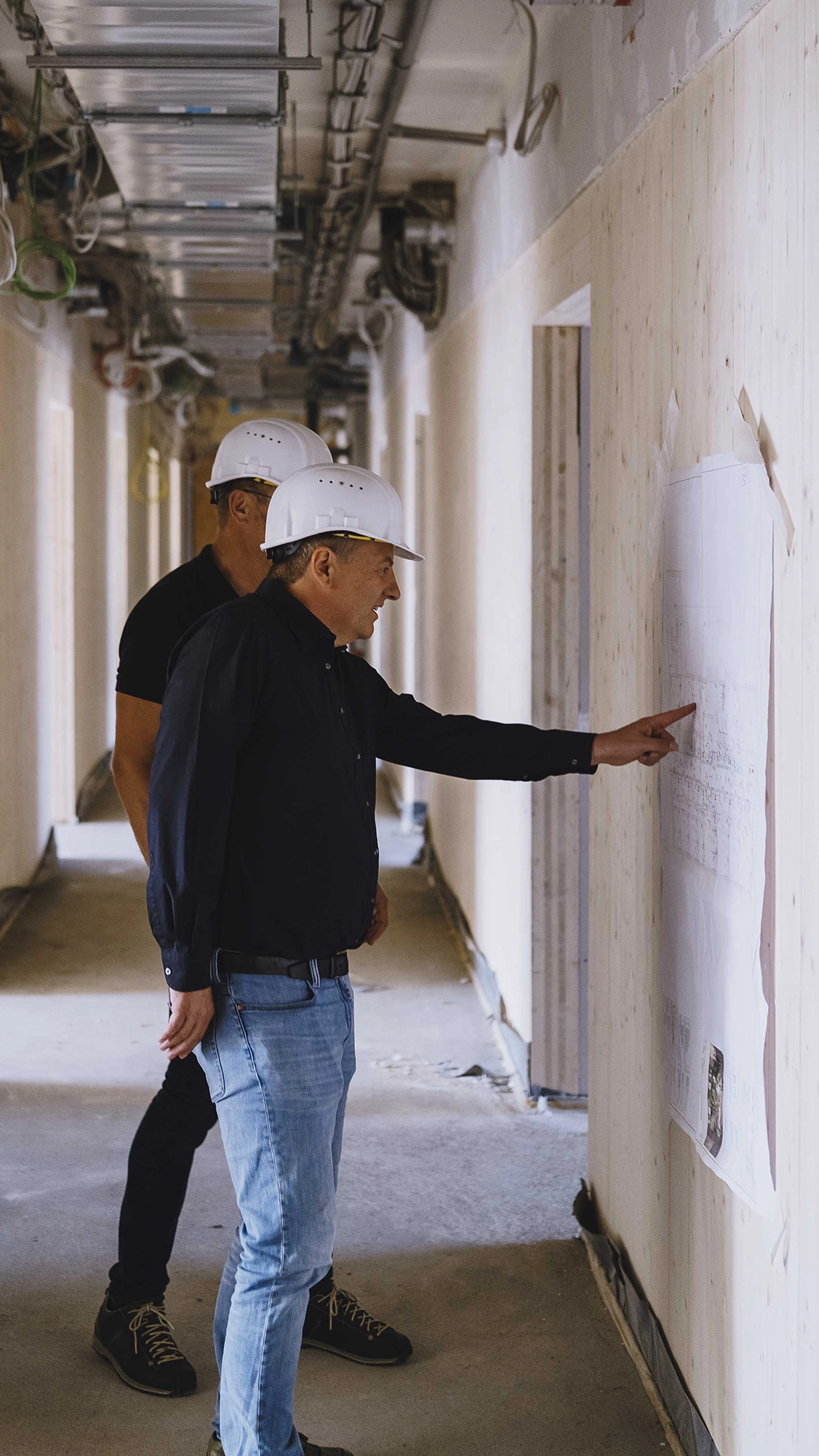 Two construction workers wearing white hard hats inspecting blueprints on a wall inside a building under construction.