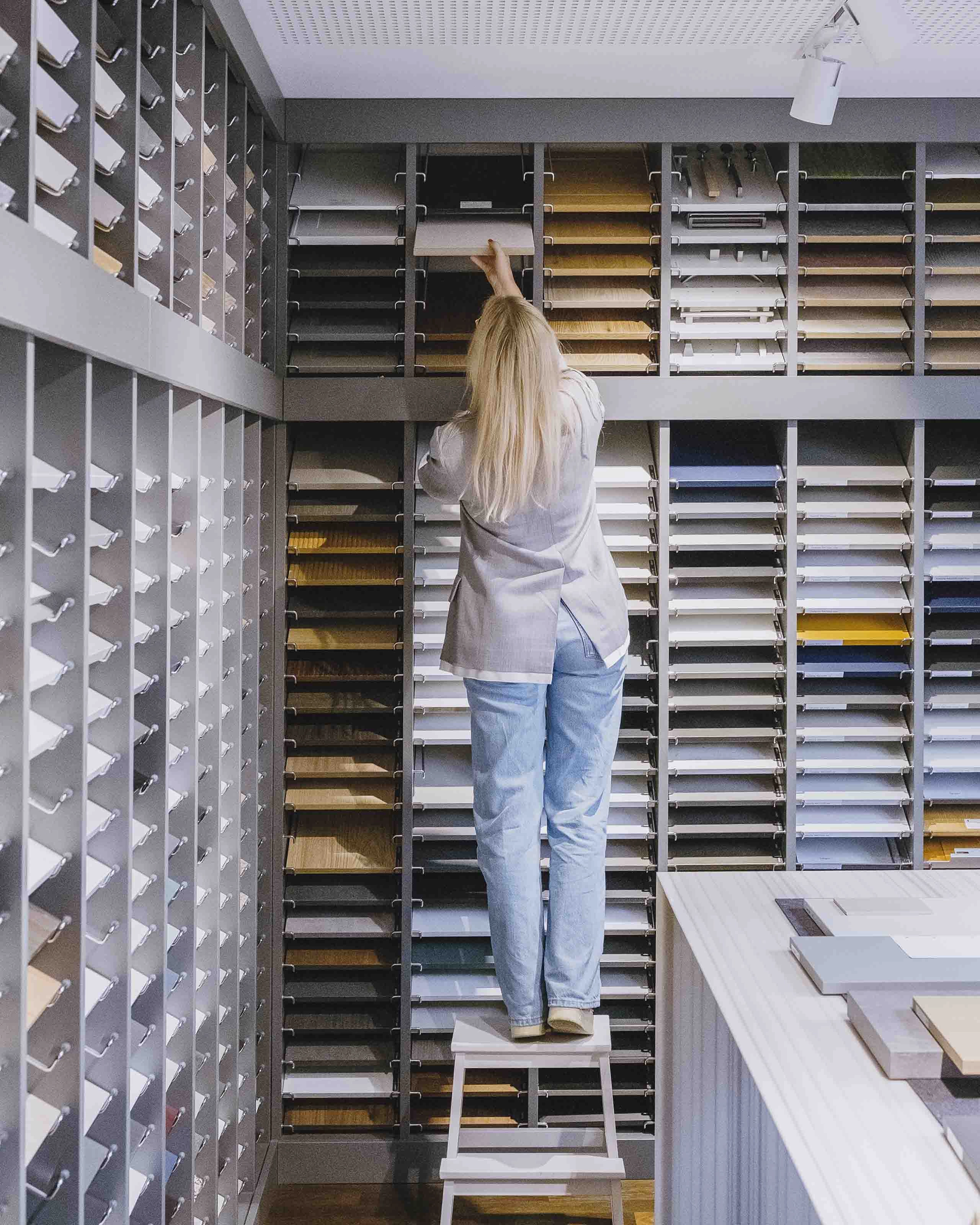 Person standing on a step stool selecting a sample from a wall-mounted display of various wood and material swatches in a showroom.