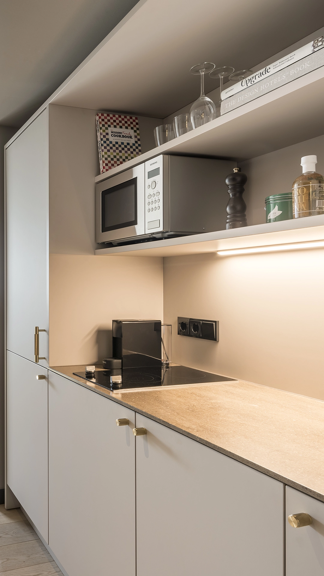 Modern kitchen corner with a microwave, stovetop, coffee machine, glassware, cookbooks, and a lighted beige countertop.