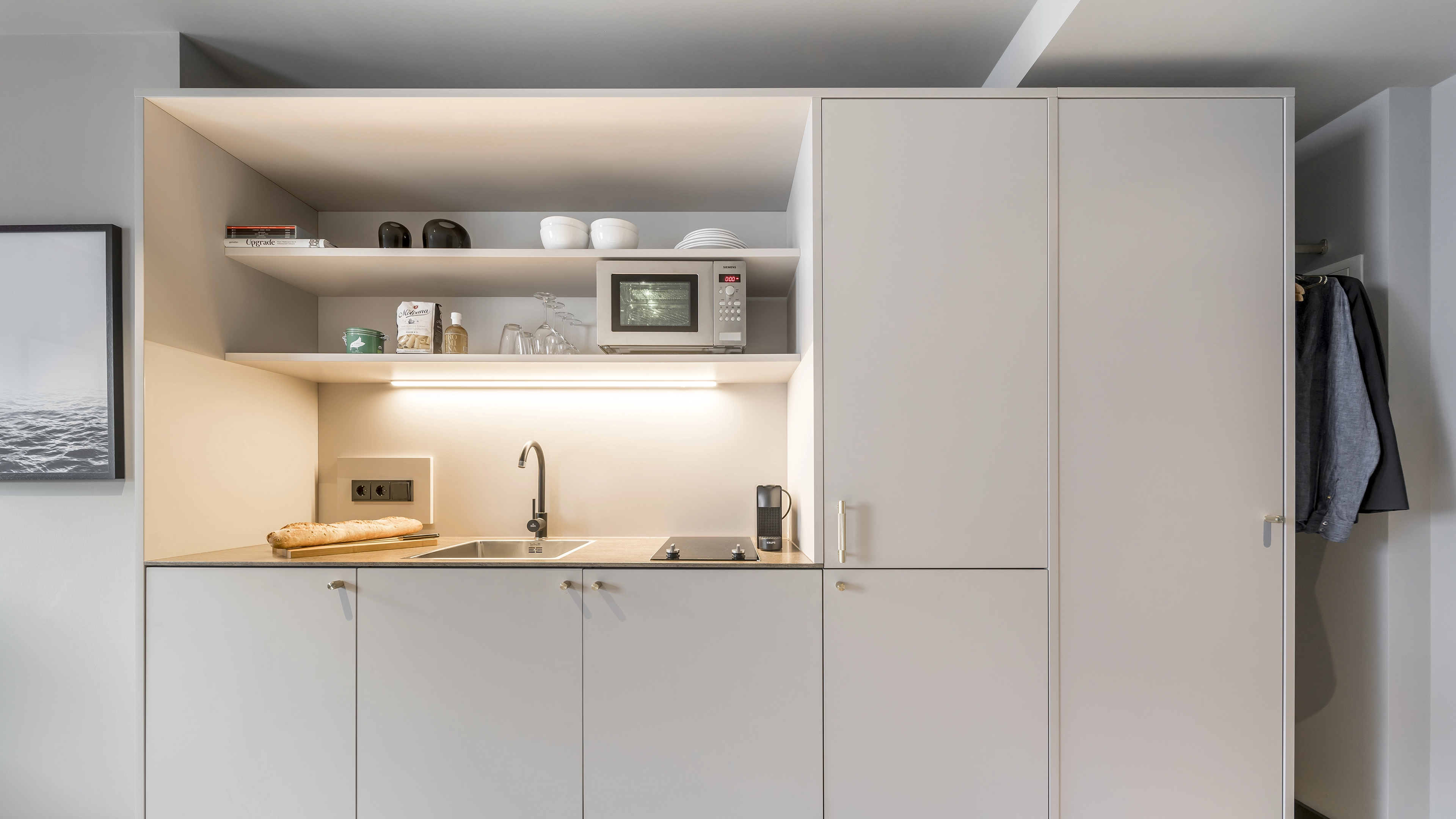 Modern compact kitchen with beige cabinets, a microwave, sink, stovetop, and a baguette on the counter.