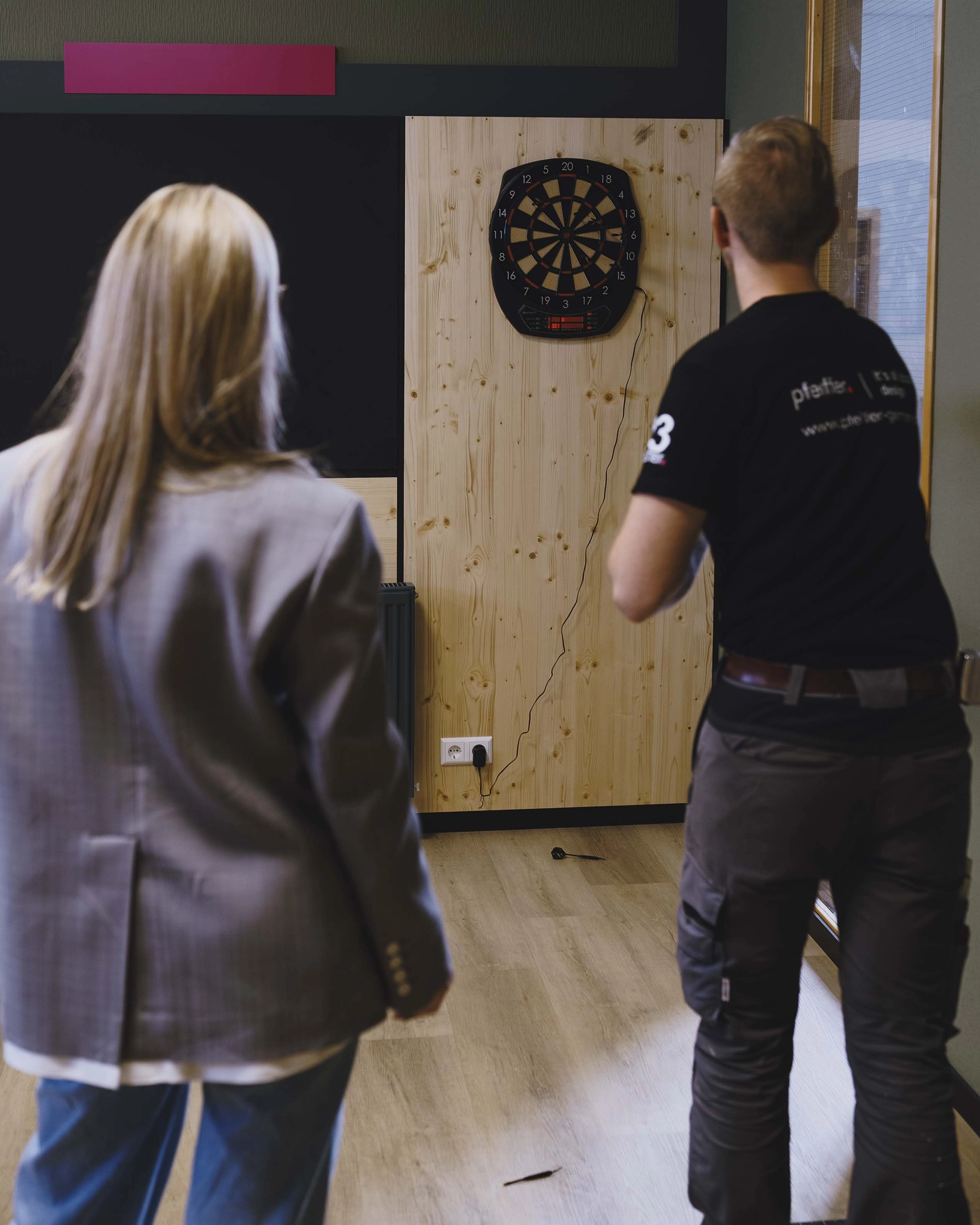Two people playing darts indoors, aiming at an electronic dartboard mounted on a wooden panel.