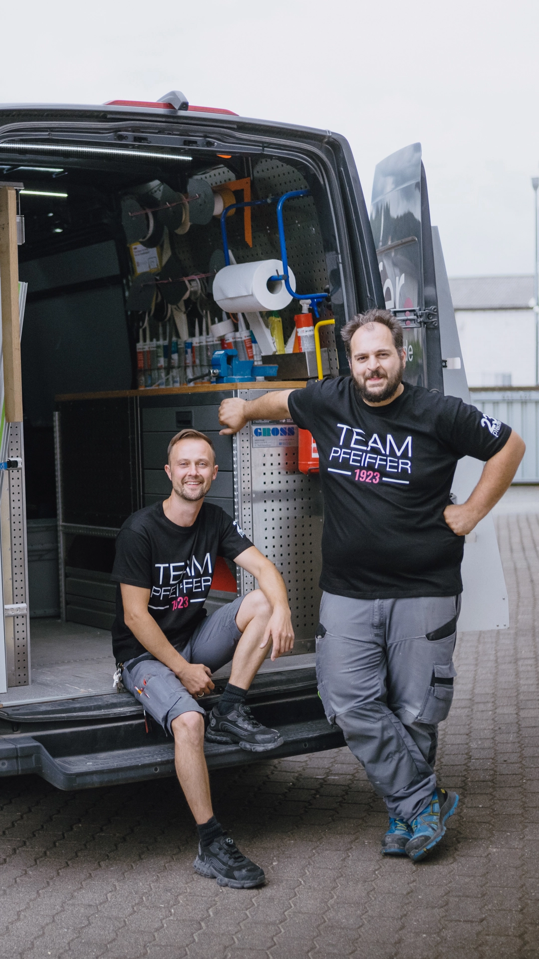 Two men wearing black Team Pfeiffer 1923 shirts standing and sitting by an open van loaded with tools.