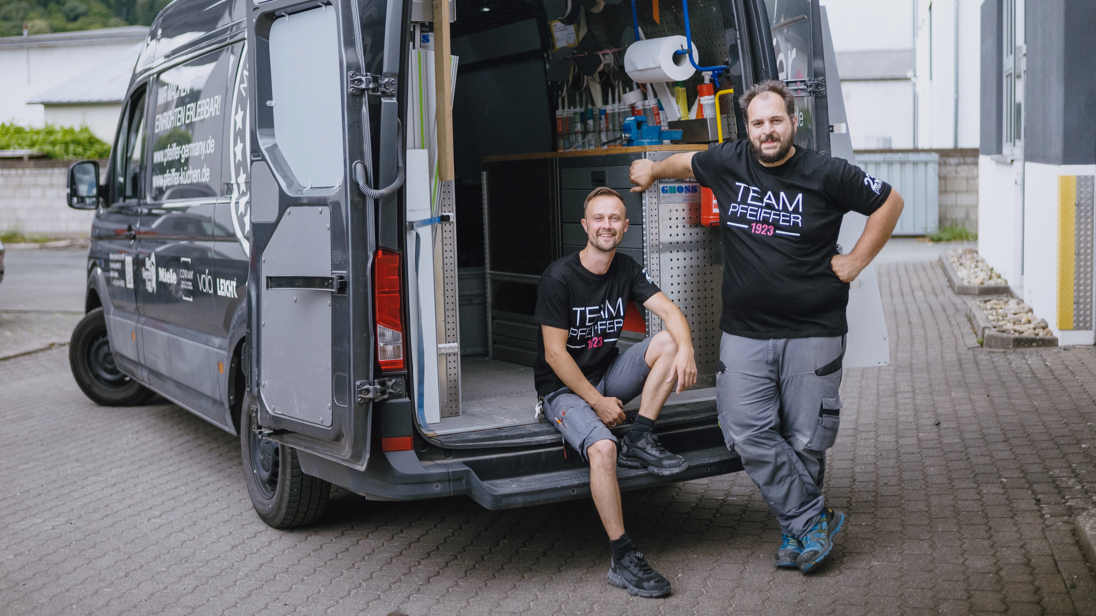 Two men wearing black Team Pfeiffer 1923 t-shirts posing by the open rear of a gray service van with tools inside.
