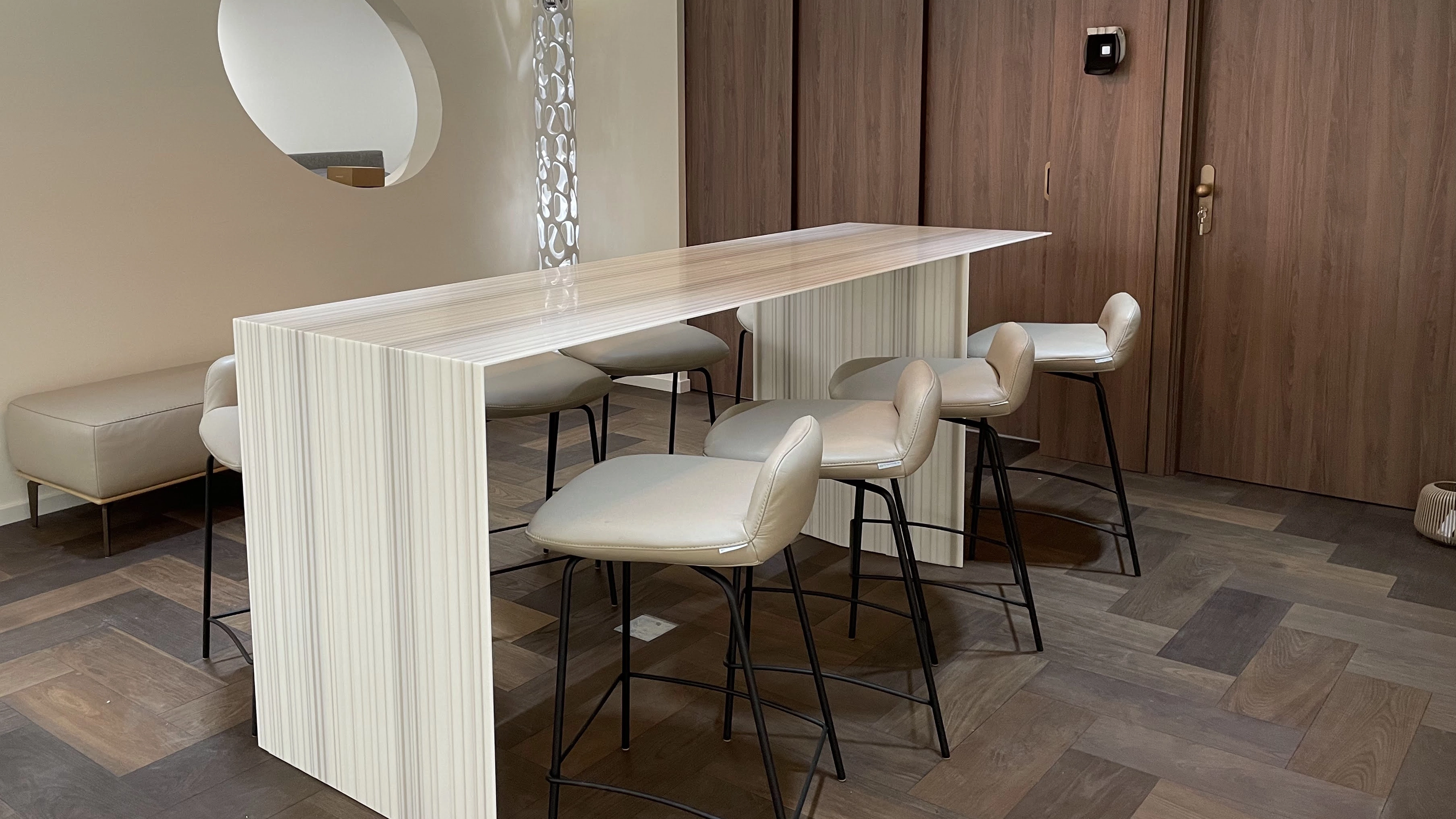 Modern dining area with a sleek, striped beige table and six cushioned beige stools on black metal legs on a patterned wooden floor.