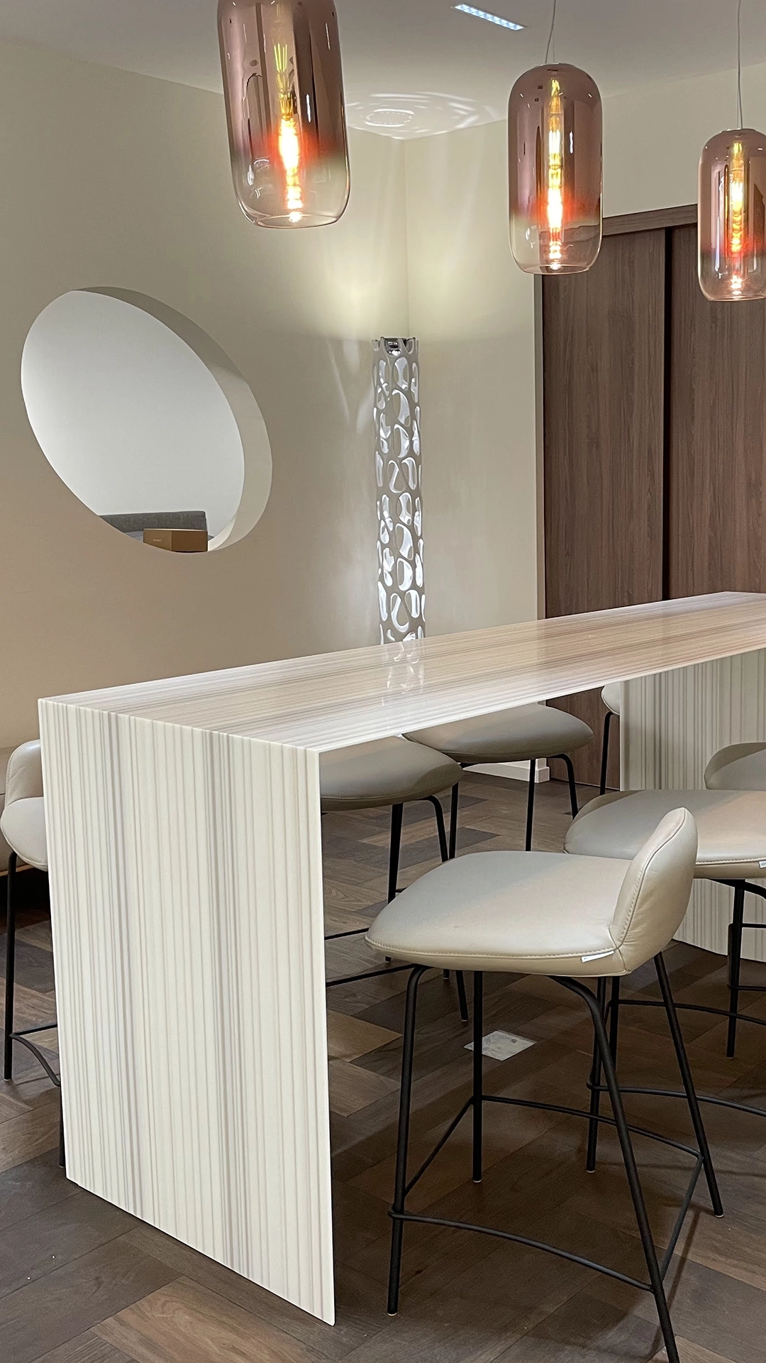 Modern dining area with a glossy white rectangular table and beige cushioned stools with black metal legs under three pendant lights with amber glass shades.
