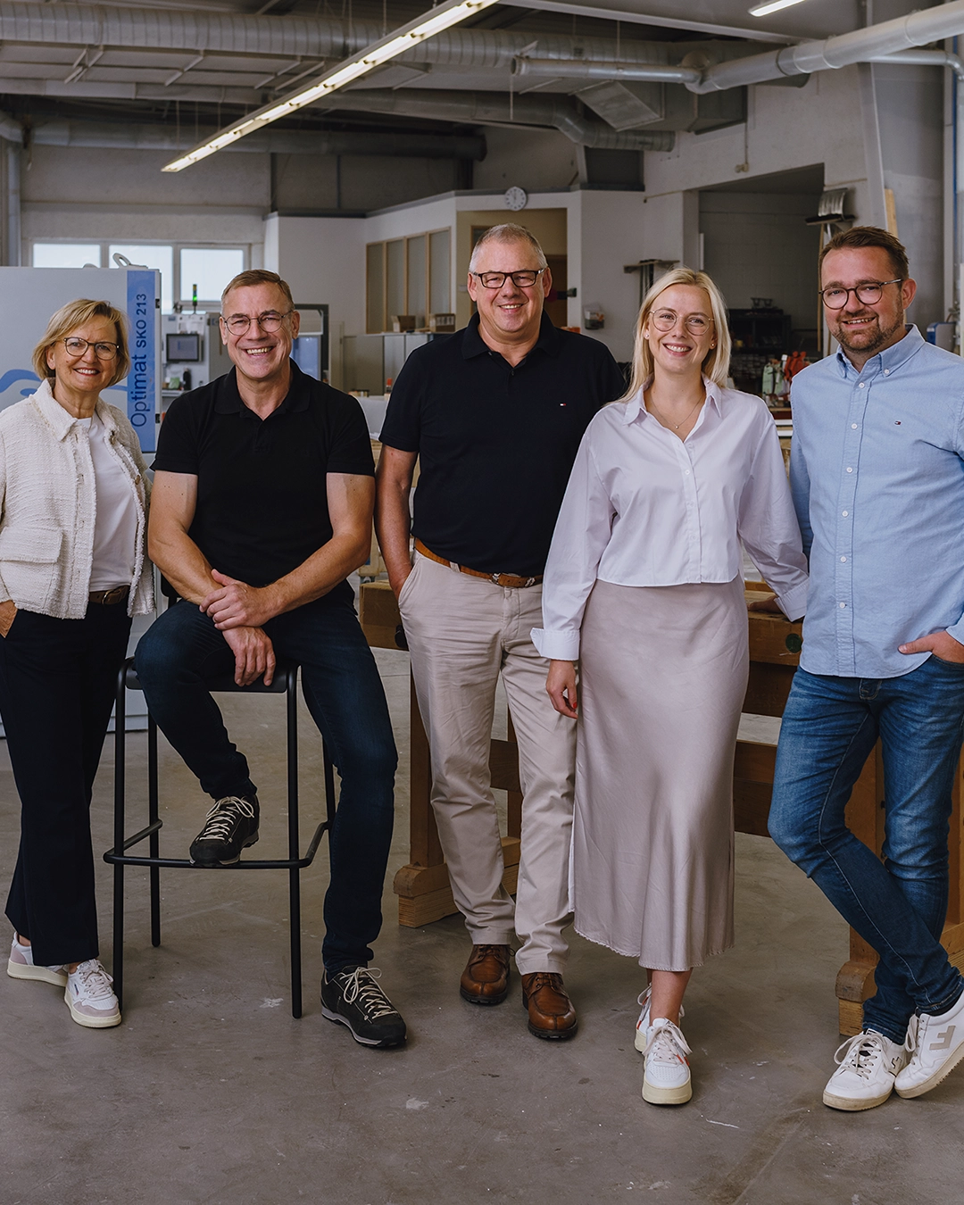 Five smiling adults wearing glasses standing and sitting in a bright industrial workspace.