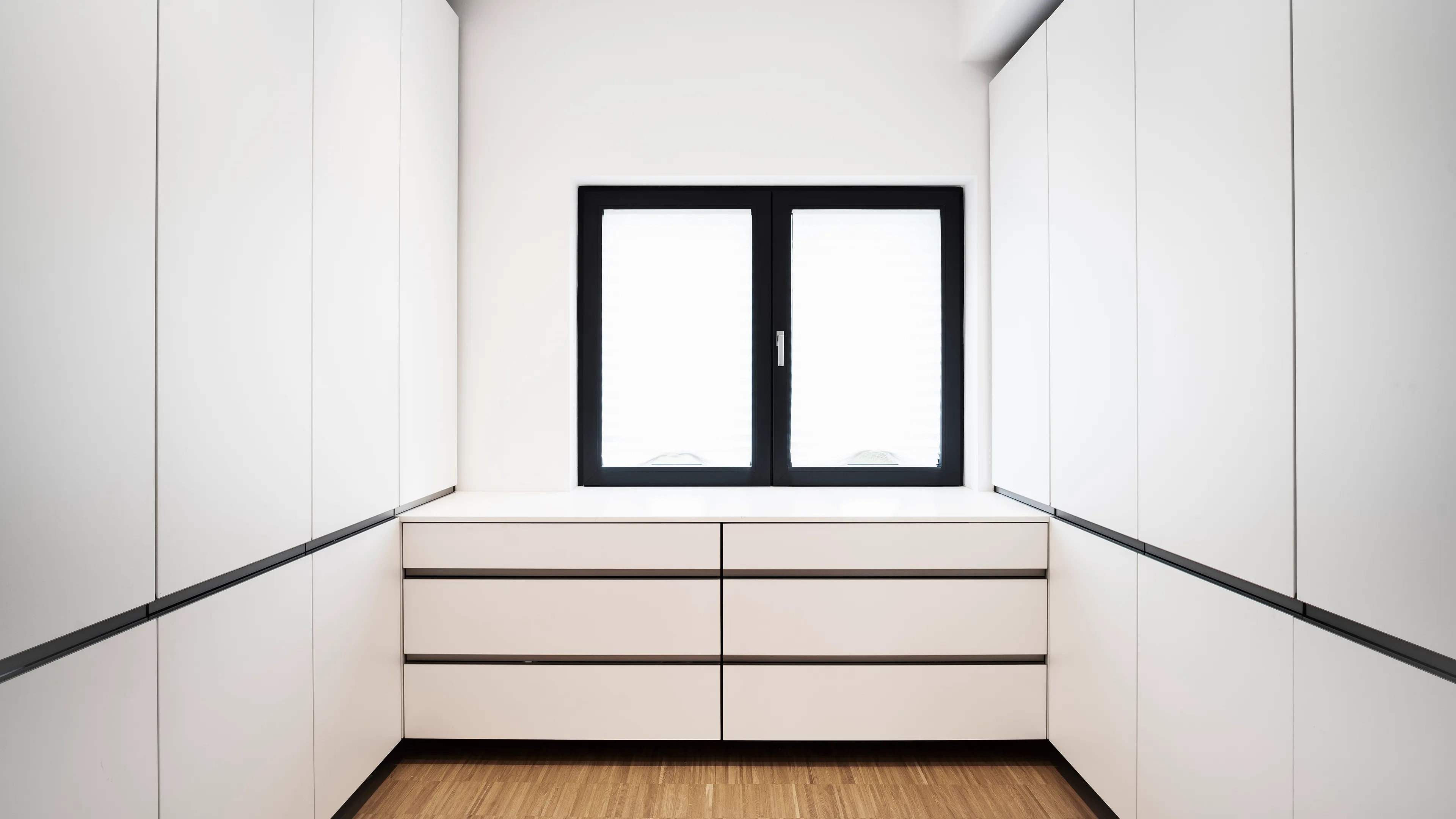 Modern minimalist white built-in cabinets with drawers below a black-framed window and wooden floor.