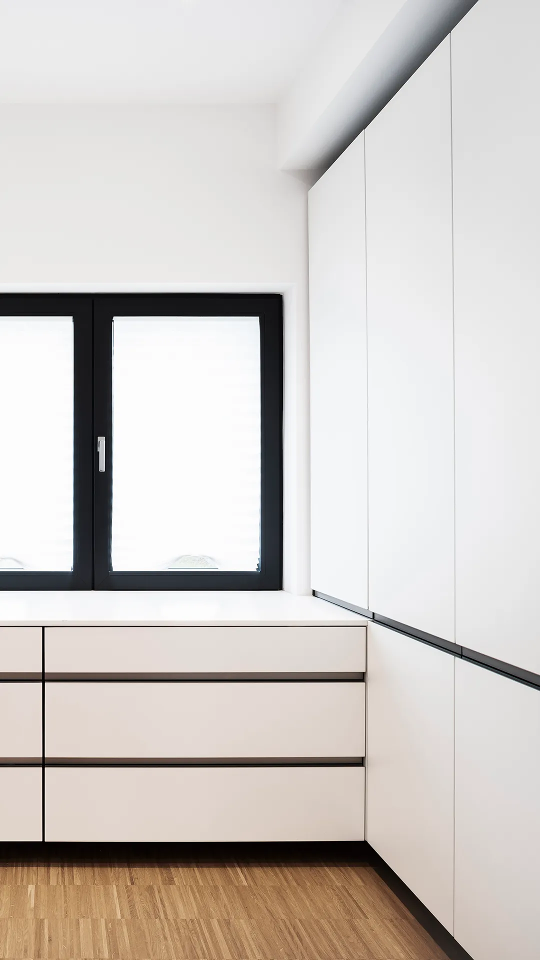 Minimalist white built-in cabinetry with drawers and clean black trim below a black-framed window with frosted glass, set against a light wooden floor.