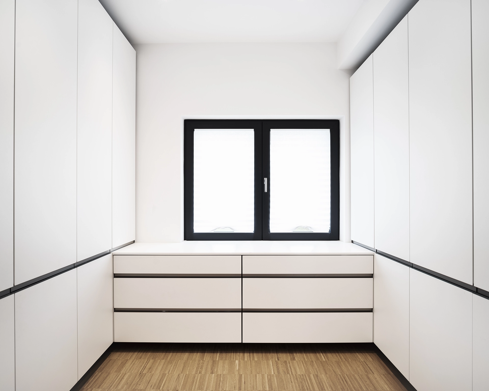 Minimalist white built-in cabinets surrounding a window with black frame and wooden floor.