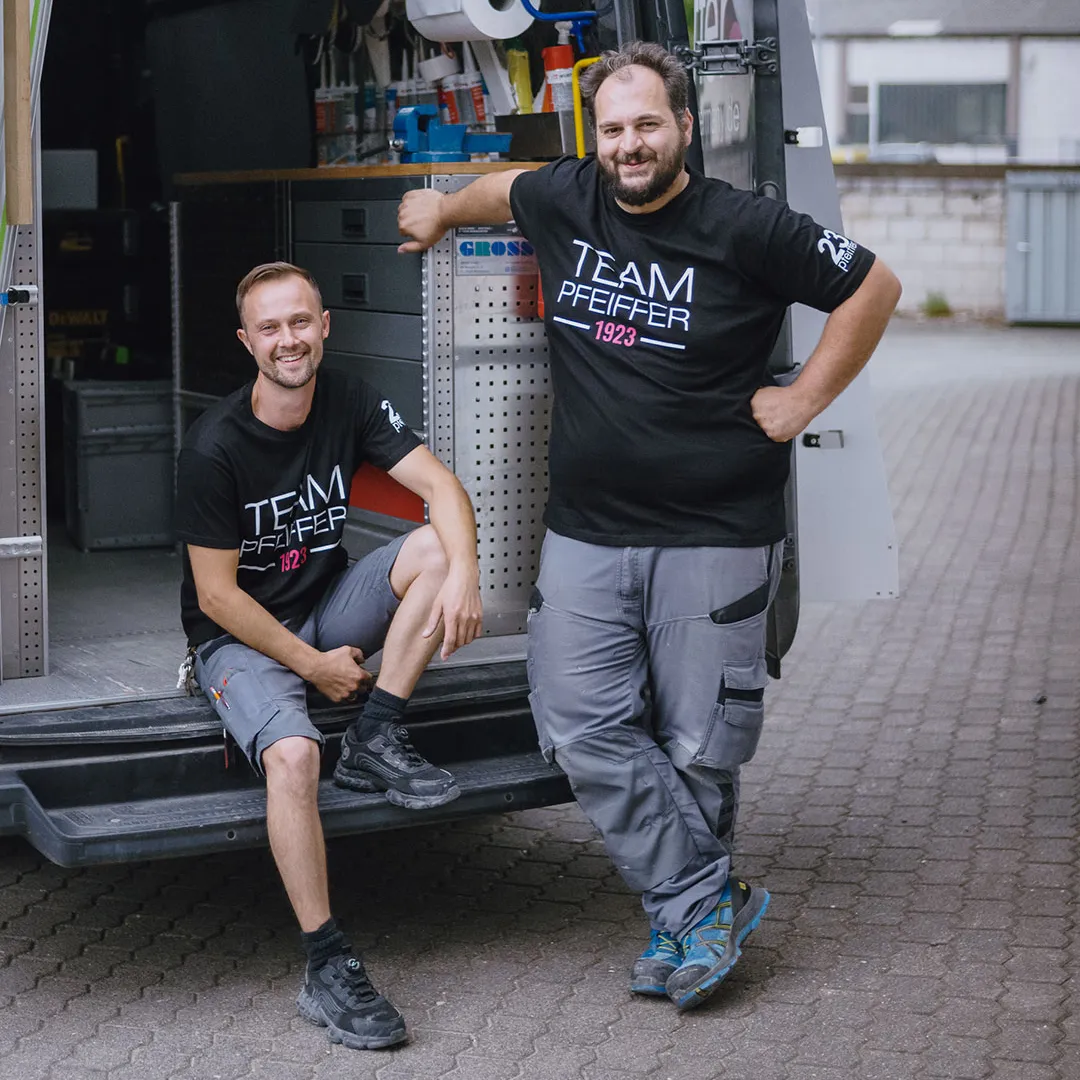 Two men in black 'Team Pfeiffer 1923' shirts smiling, one sitting on a van step and the other leaning against the van.