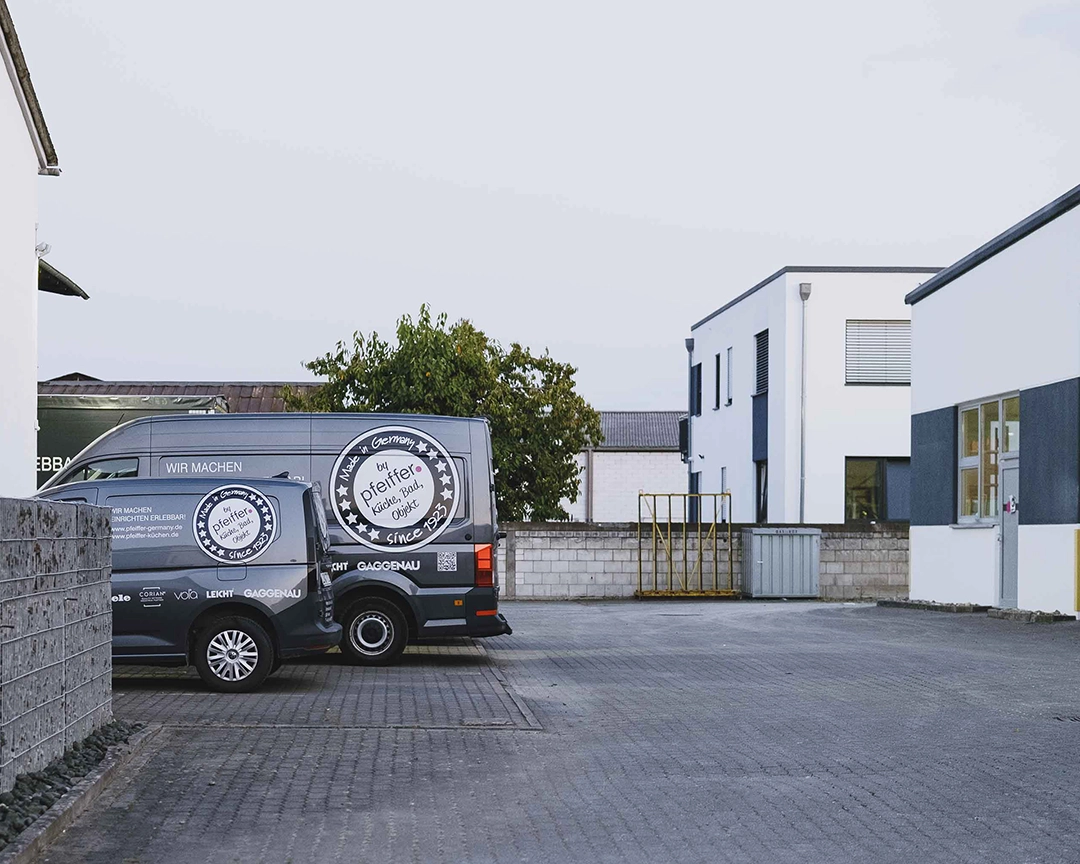 Two gray company vans with Pfeiffer Kitchens branding parked in an industrial lot with white buildings and a brick wall in the background.