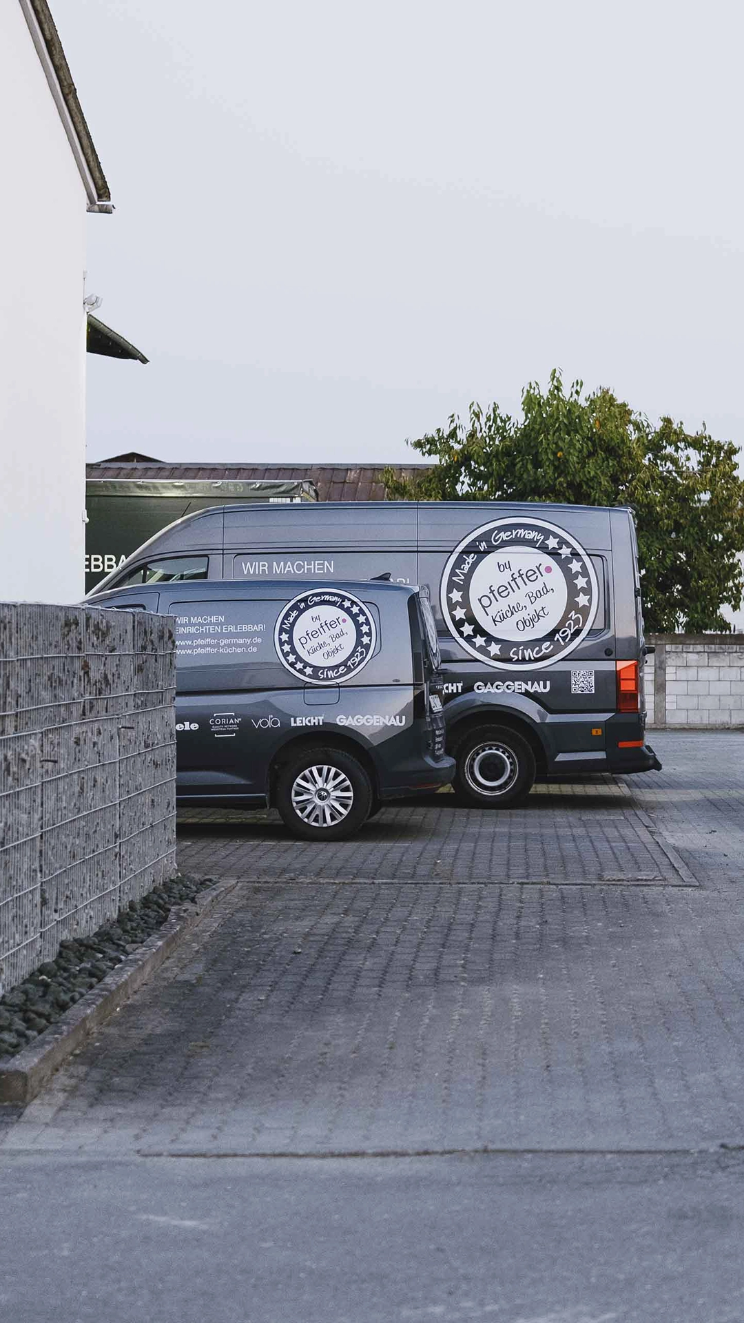 Two dark blue vans parked side by side on a paved lot, each with circular white logos and text advertising Pfeiffer kitchens and bathrooms.