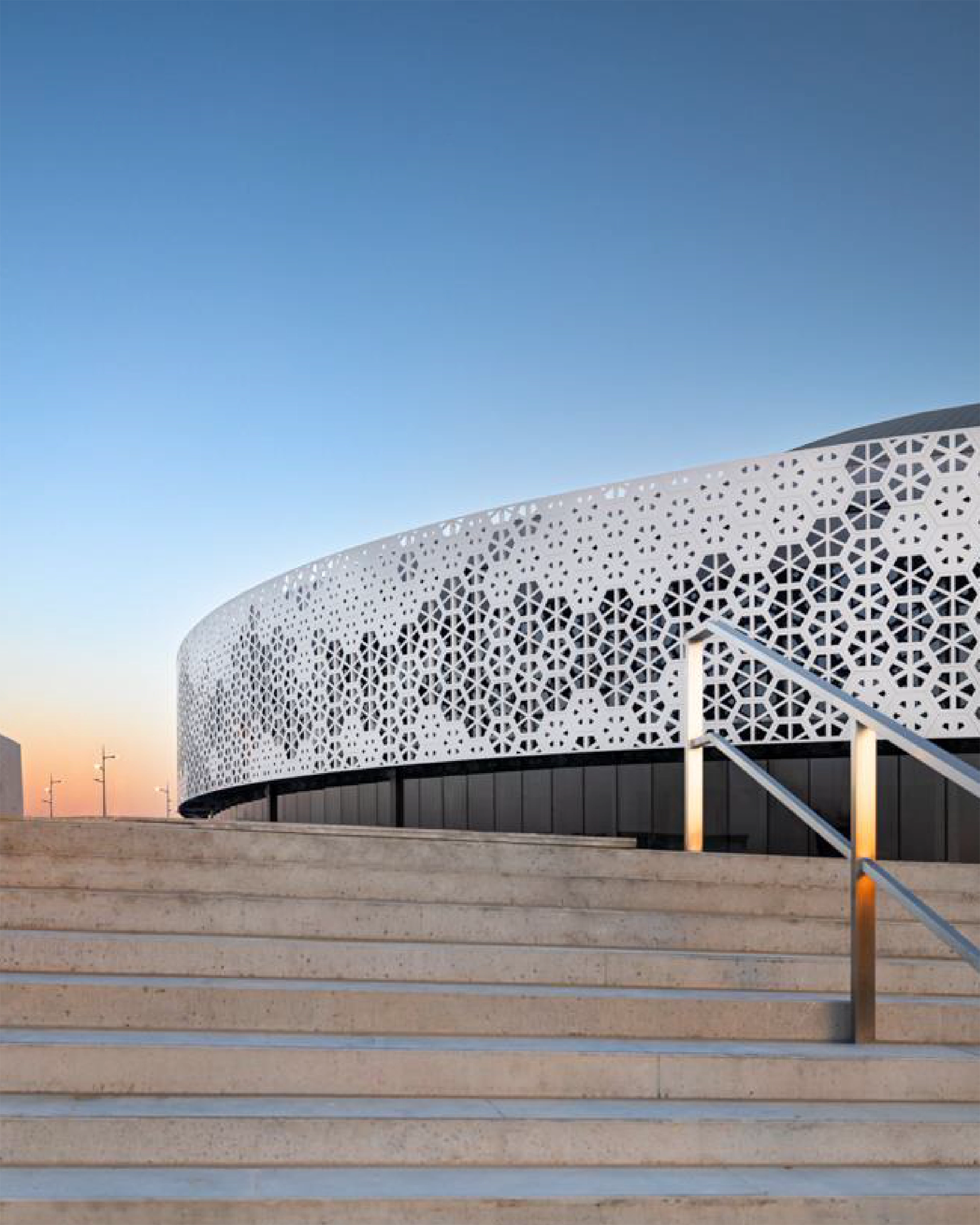 Wide stone staircase leading up to a modern building with a perforated white patterned metal facade under a clear blue sky.