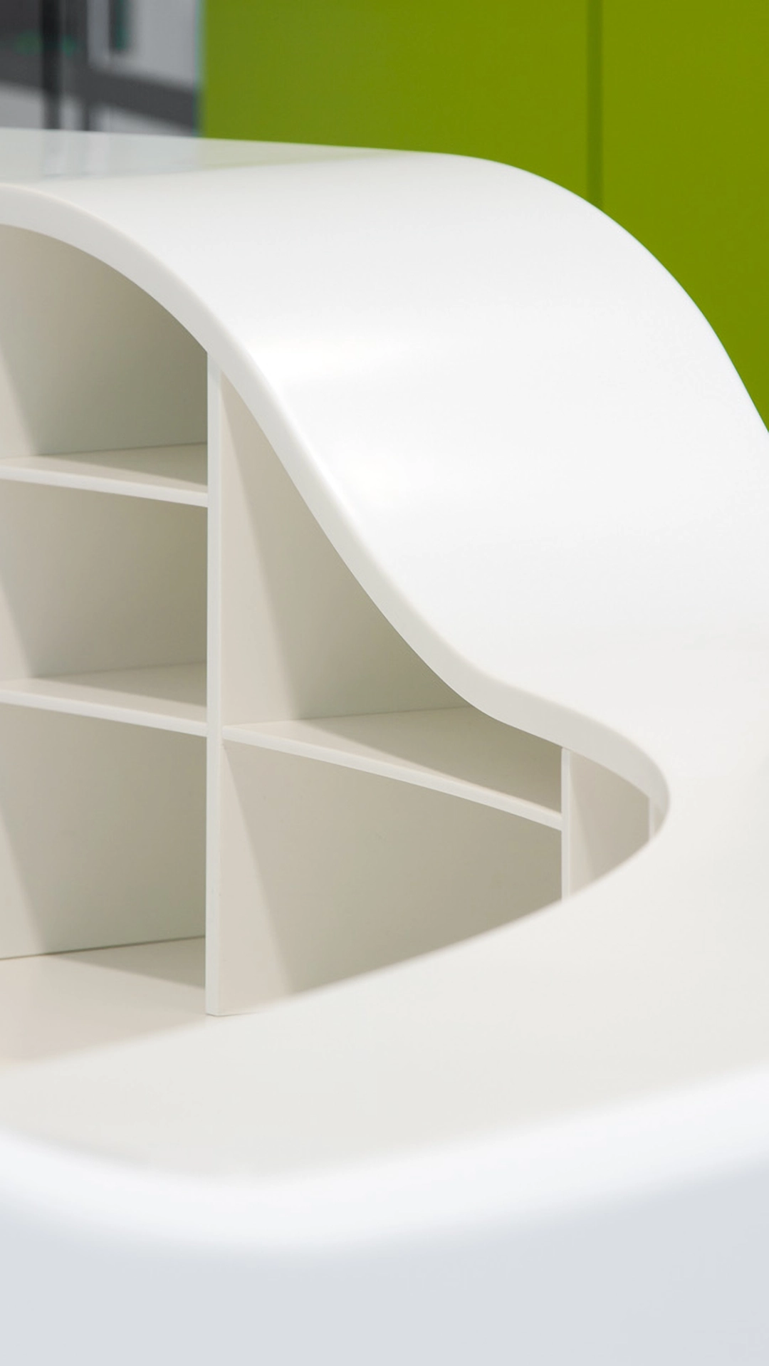 Close-up of a modern white curved shelf unit with multiple compartments against a green wall.