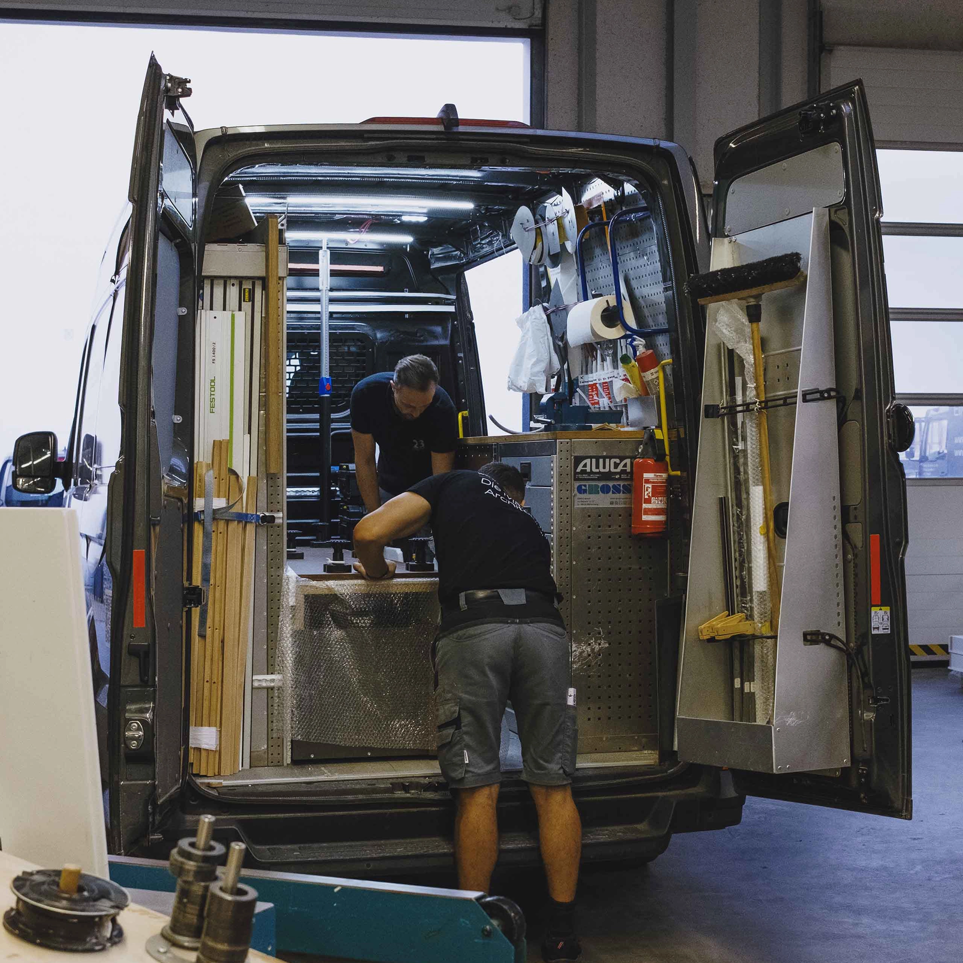 Zwei Männer laden Werkzeuge und Materialien aus einem Lieferwagen in einer Werkstatt aus.