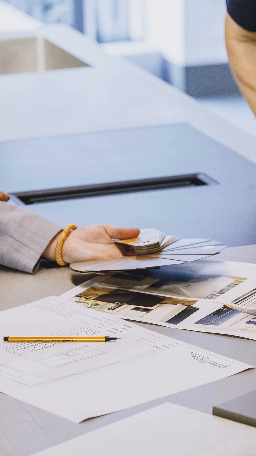 Eine Hand hält Farbfächer über einem Tisch mit Architekturplänen, Fotos und einem gelben Stift.