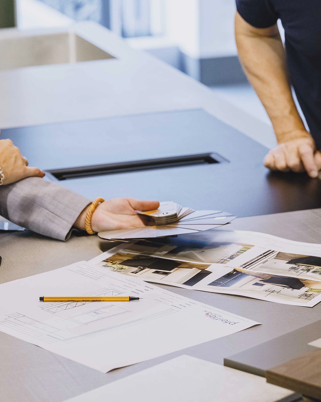 Zwei Personen diskutieren an einem Tisch mit Farbmustern, Architekturzeichnungen und Fotos von Innenräumen.