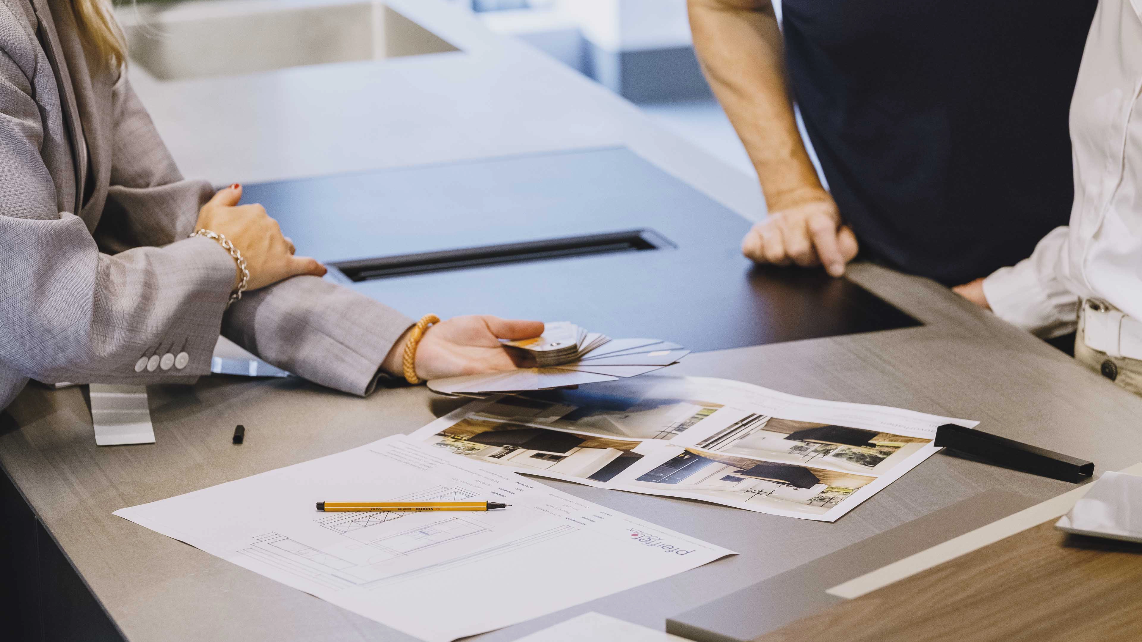 Person zeigt Farbproben und Architekturpläne auf einem Tisch mit zwei weiteren Personen in einem Büro.