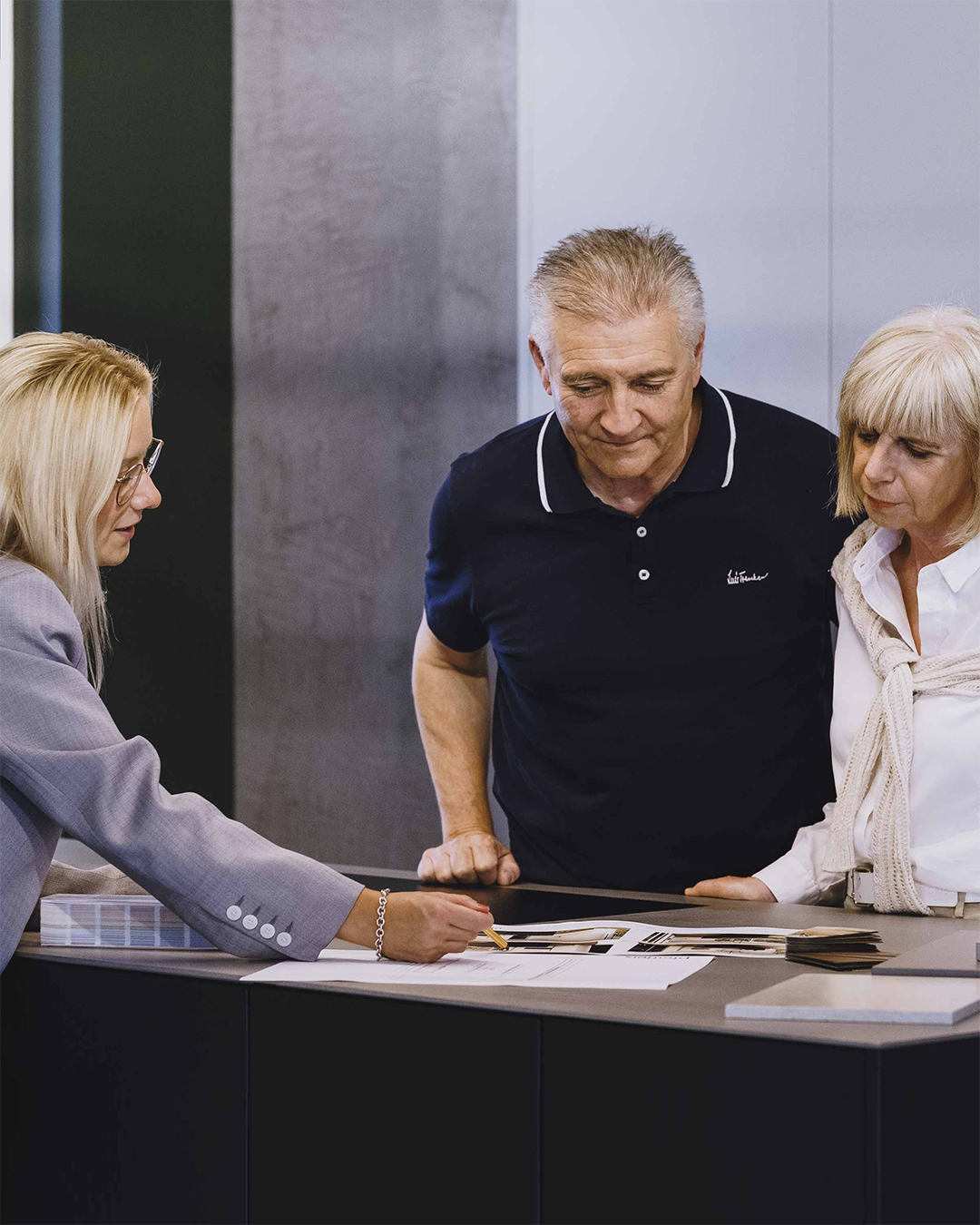 Eine Beraterin zeigt einem älteren Paar am Tresen Planungsunterlagen und Muster.