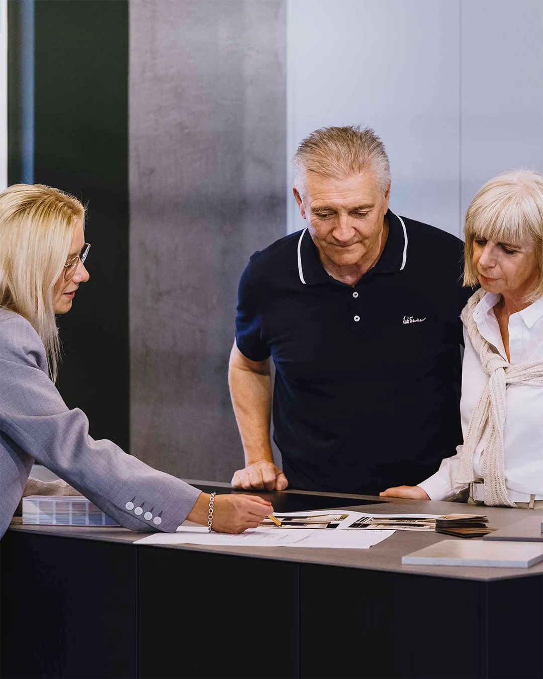 Eine Beraterin zeigt einem älteren Paar am Tresen Planungsunterlagen und Muster.