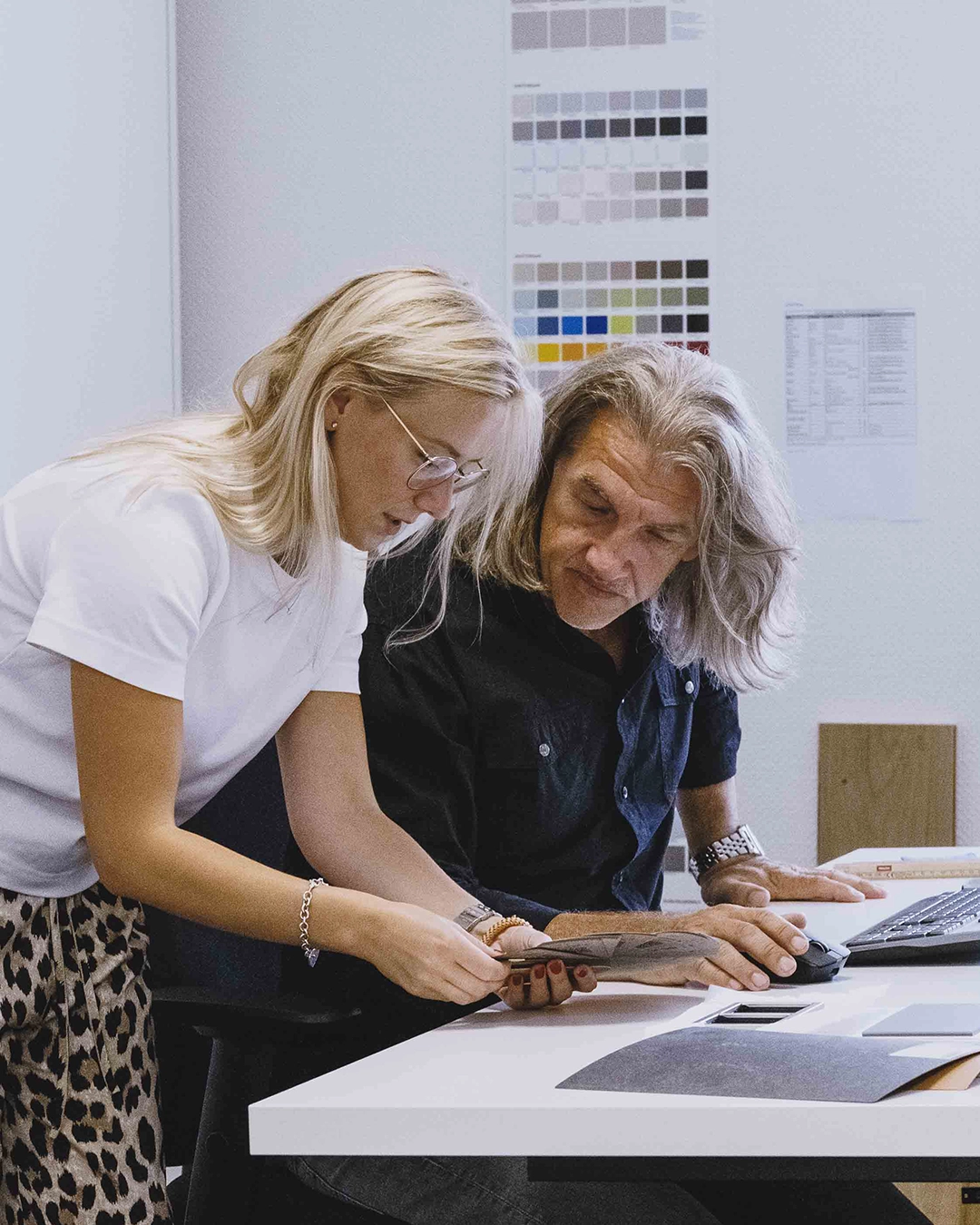 Zwei Personen diskutieren über ein Musterstück in einem Büro mit Farbmusterkarten an der Wand.