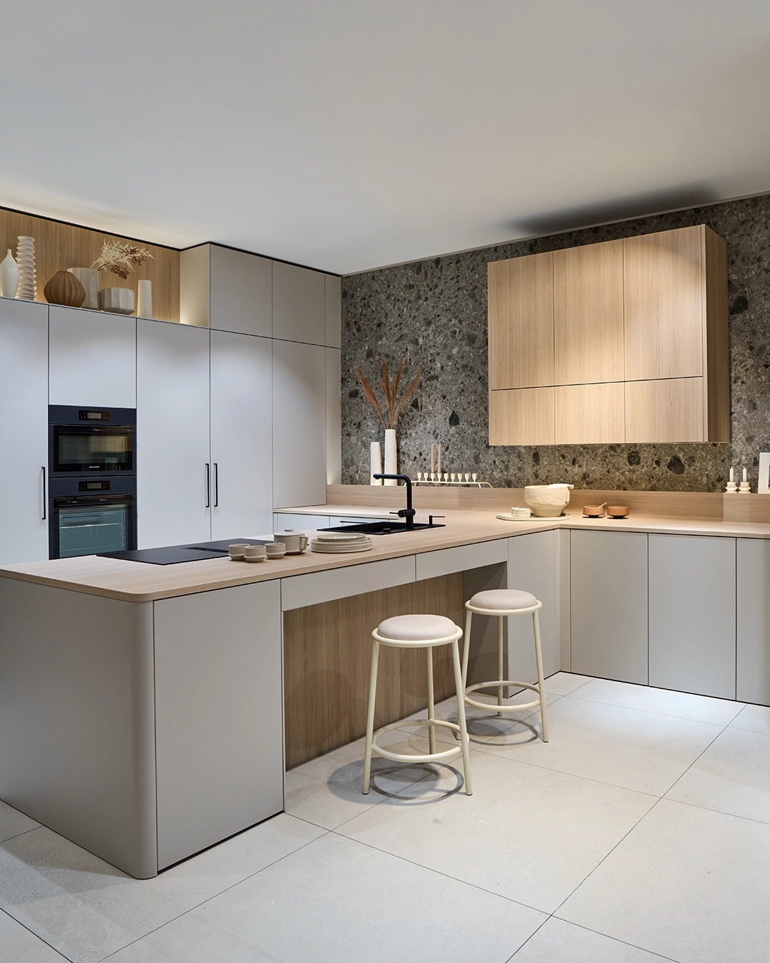 Moderne Küche mit grauen Schränken, Holzarbeitsplatte, zwei weißen Hockern und grauer Steinwand im Hintergrund.
