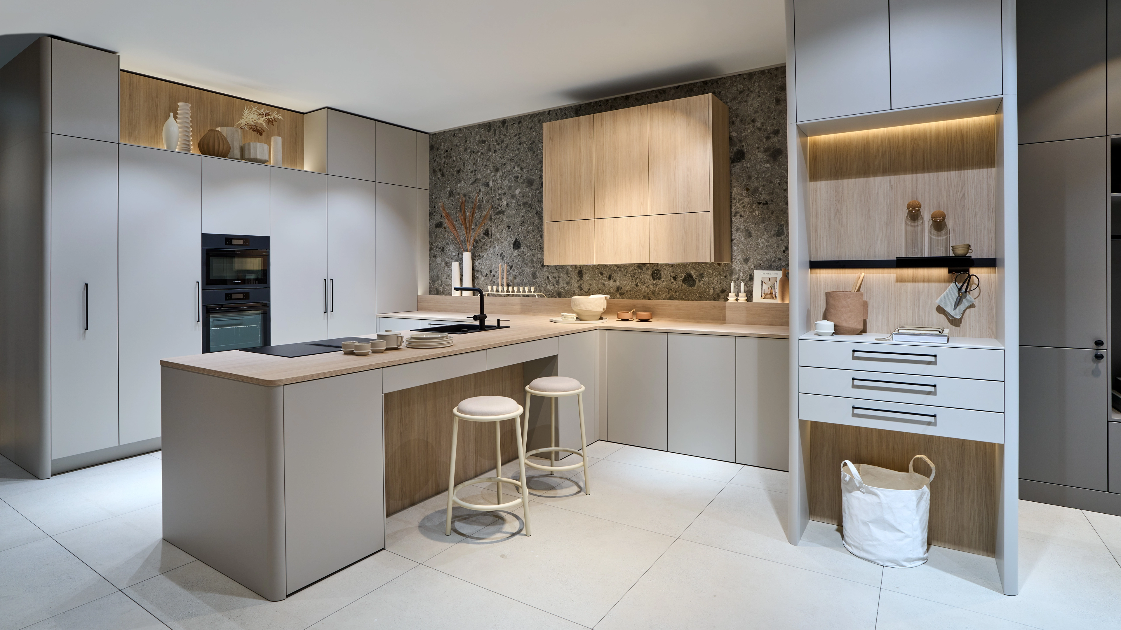 Moderne U-förmige Küche mit hellen Schränken, Holzarbeitsplatten, zwei weißen Hockern und schwarzem Wasserhahn vor grauer Steinwand.