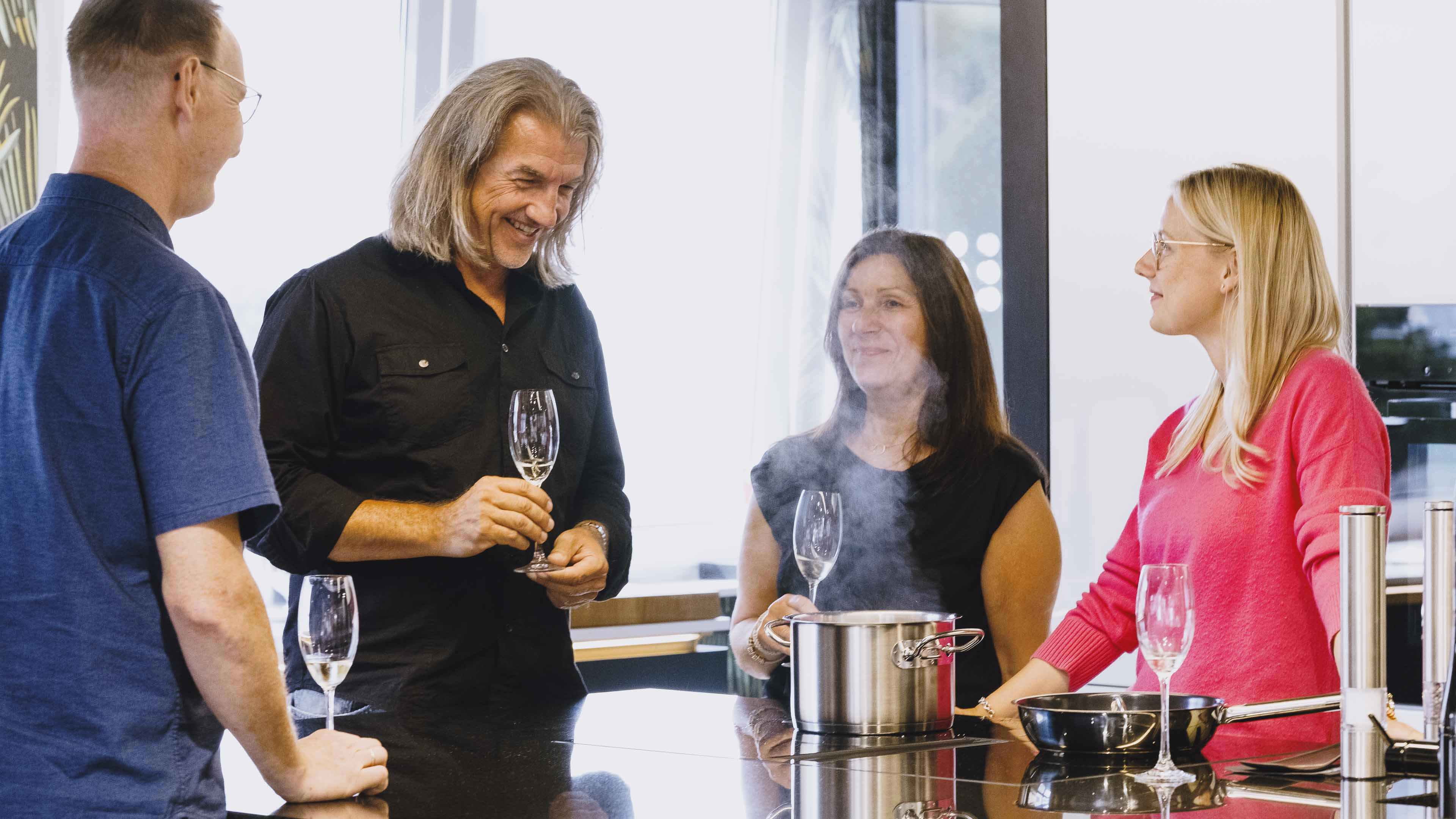 Vier Personen stehen an einem Küchentresen, unterhalten sich und halten Sektgläser, während ein Dampf aus einem Kochtopf aufsteigt.