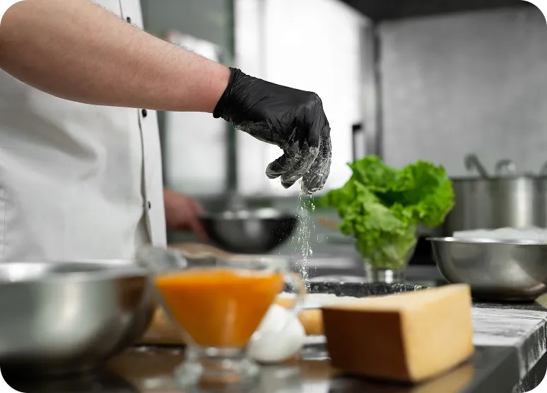 Chef wearing black glove sprinkling flour over a countertop with ingredients like lettuce, cheese, and sauce in a kitchen.