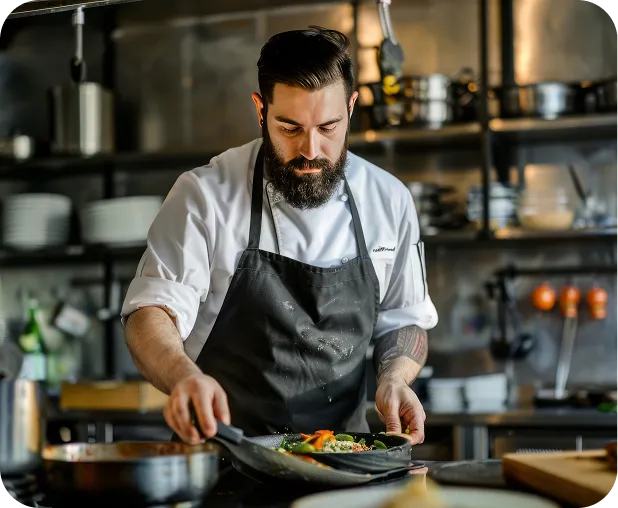 Bearded chef in black apron preparing food in a professional kitchen.