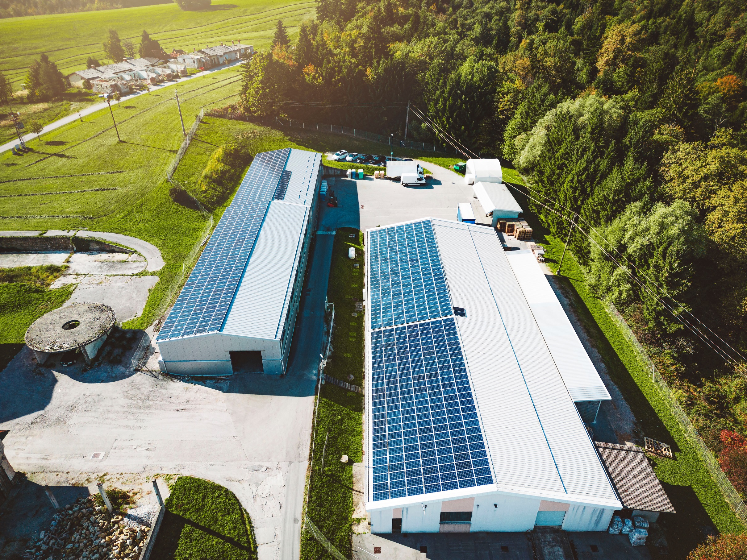 Aerial view of two industrial buildings with solar panels on the roofs surrounded by green fields and trees.