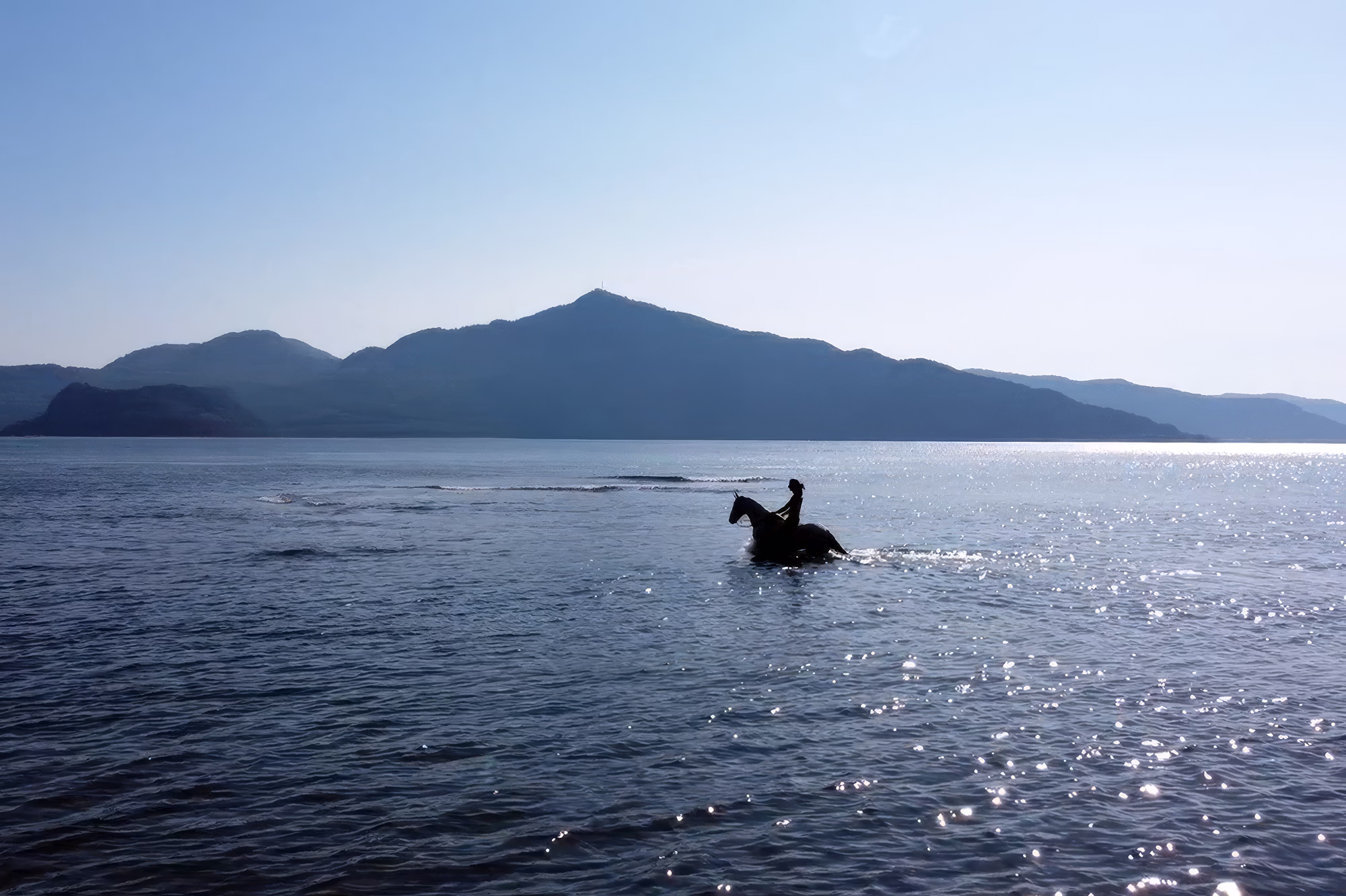 A woman riding a horse through deep water with scenic hills in the background