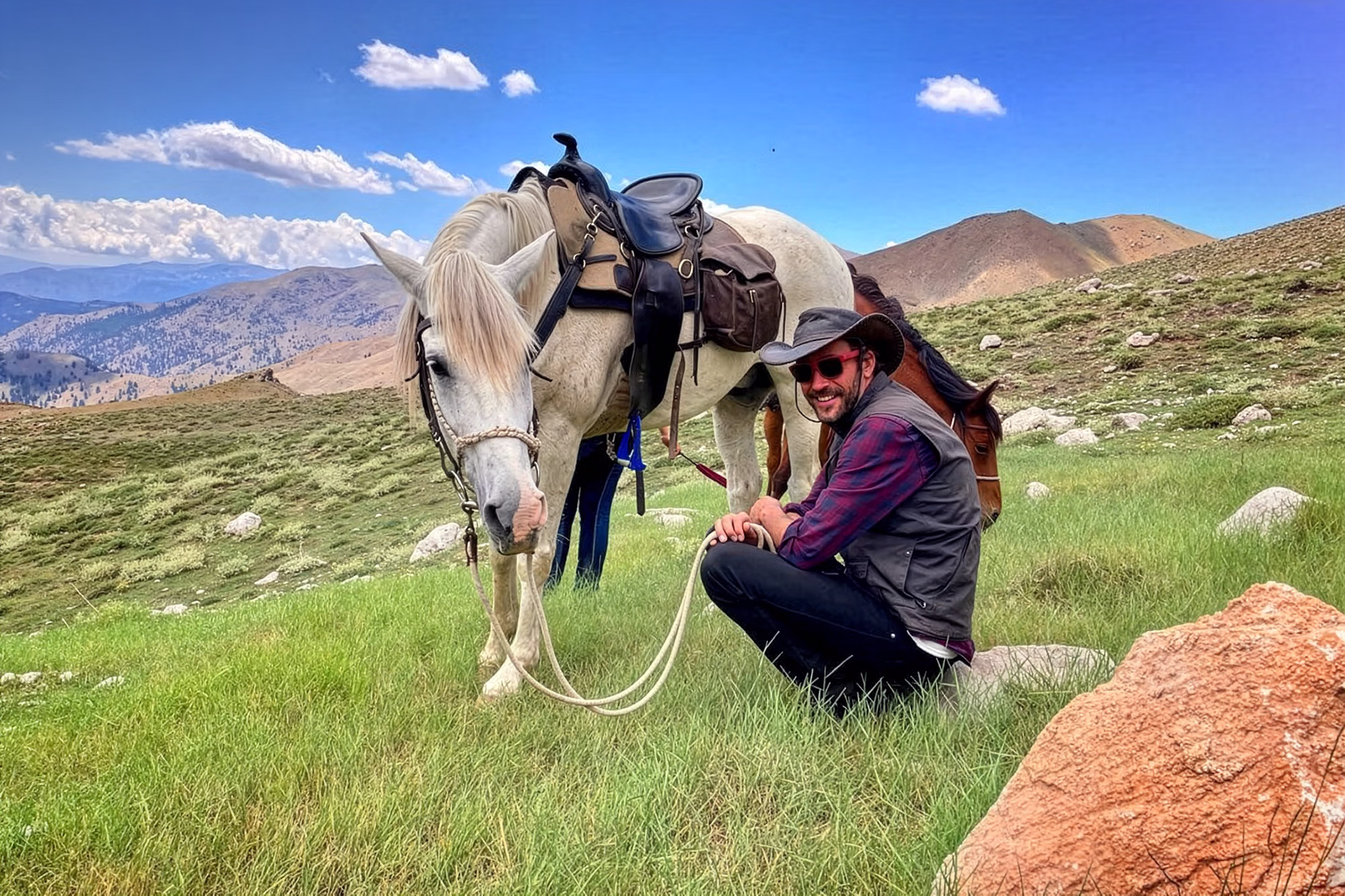A smiling rider wearing a hat is crouching in a grassy landscape while holding the lead rope of their horse