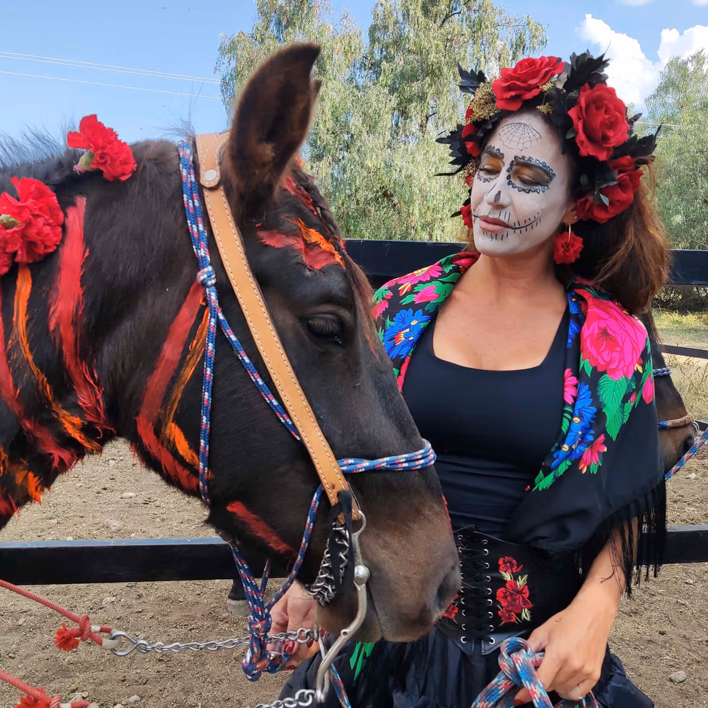 A woman in full Día de los Muertos attire standing with a trail horse decorated with red paint and flowers