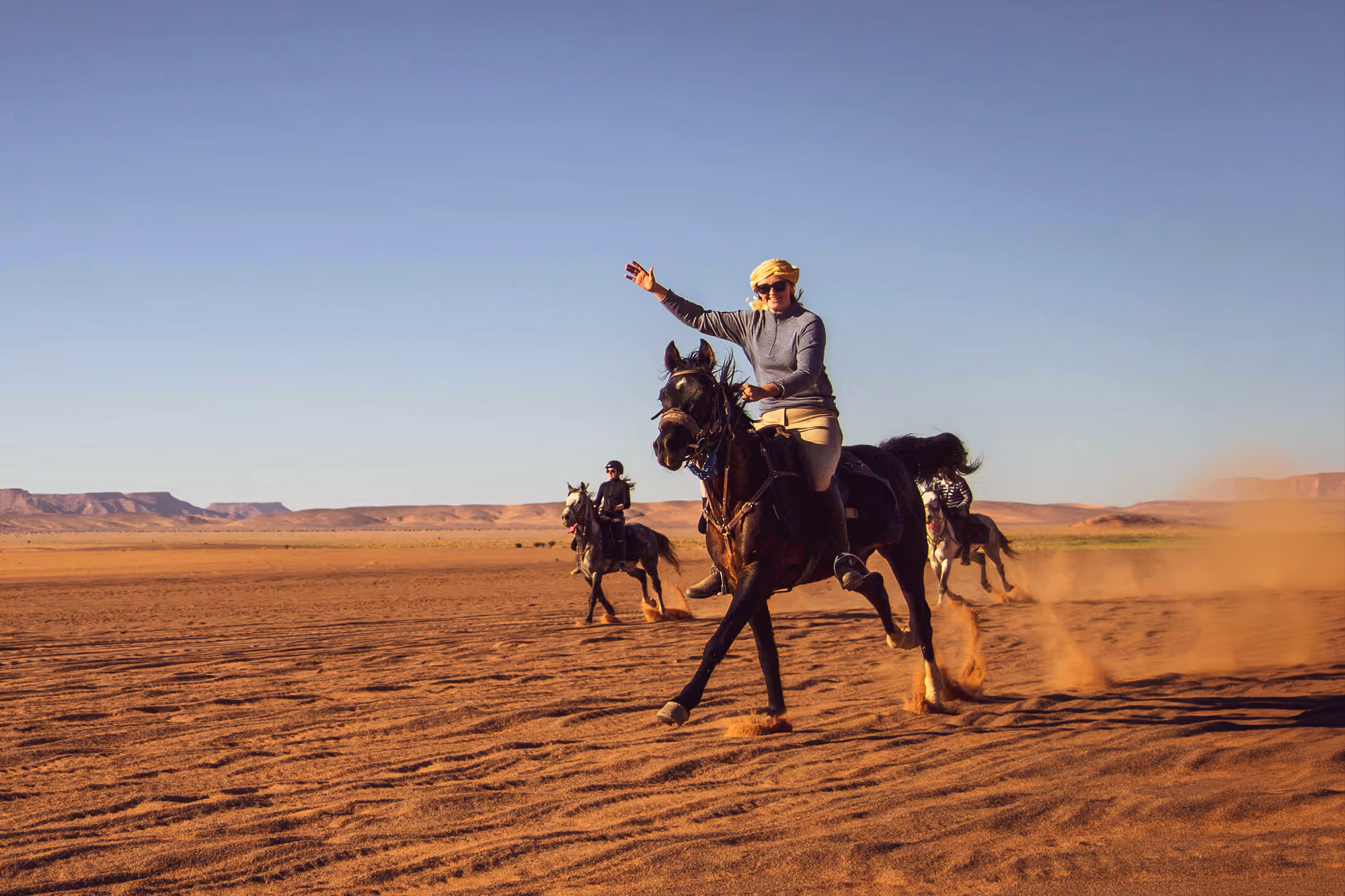 A smiling woman in a headscarf waves while galloping on her horse ahead of a group in the Merzouga desert dunes.