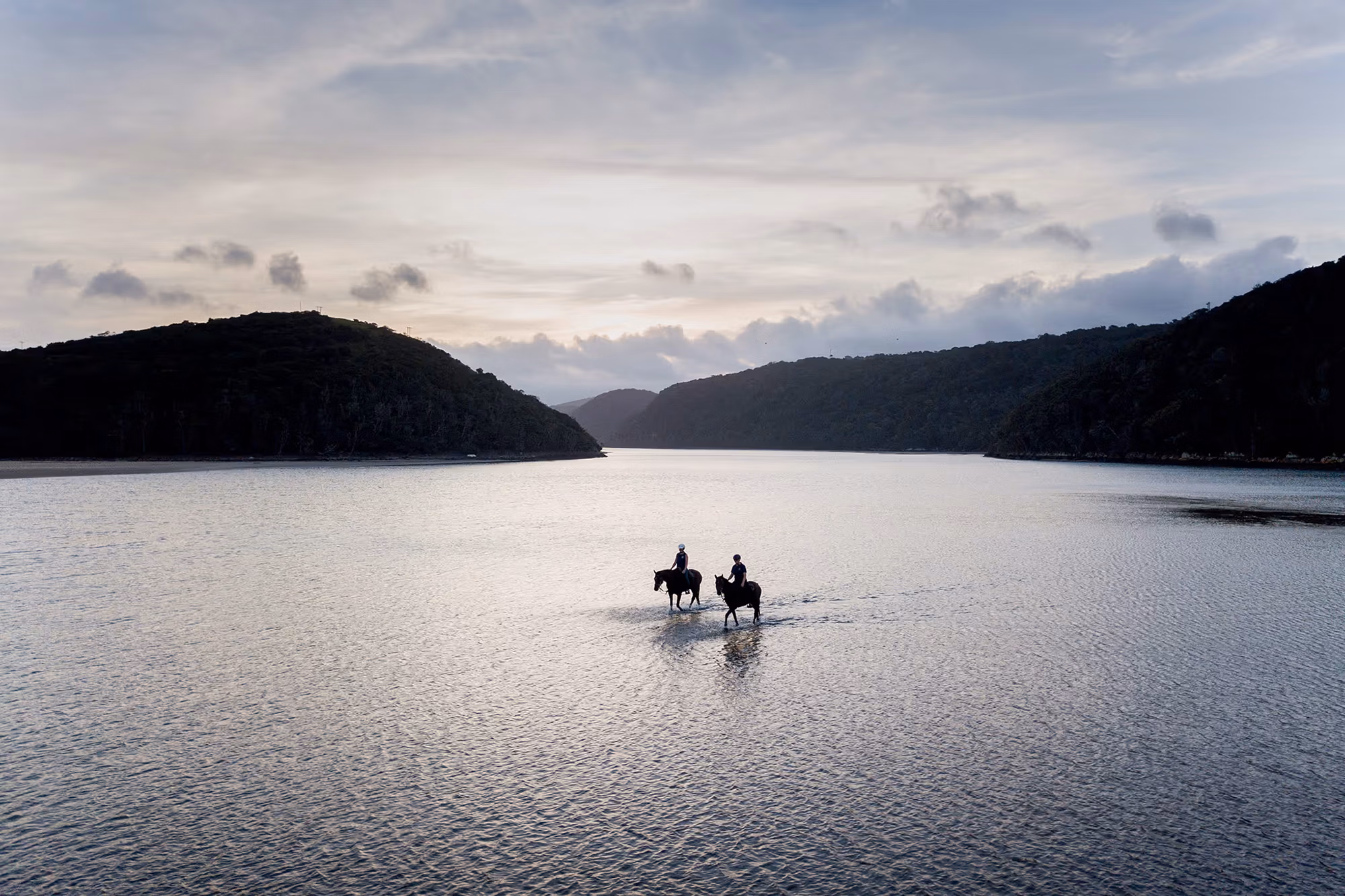 A bird's-eye panoramic view of two equestrians riding through coastal waters on the Eastern Cape horse trail in South Africa.