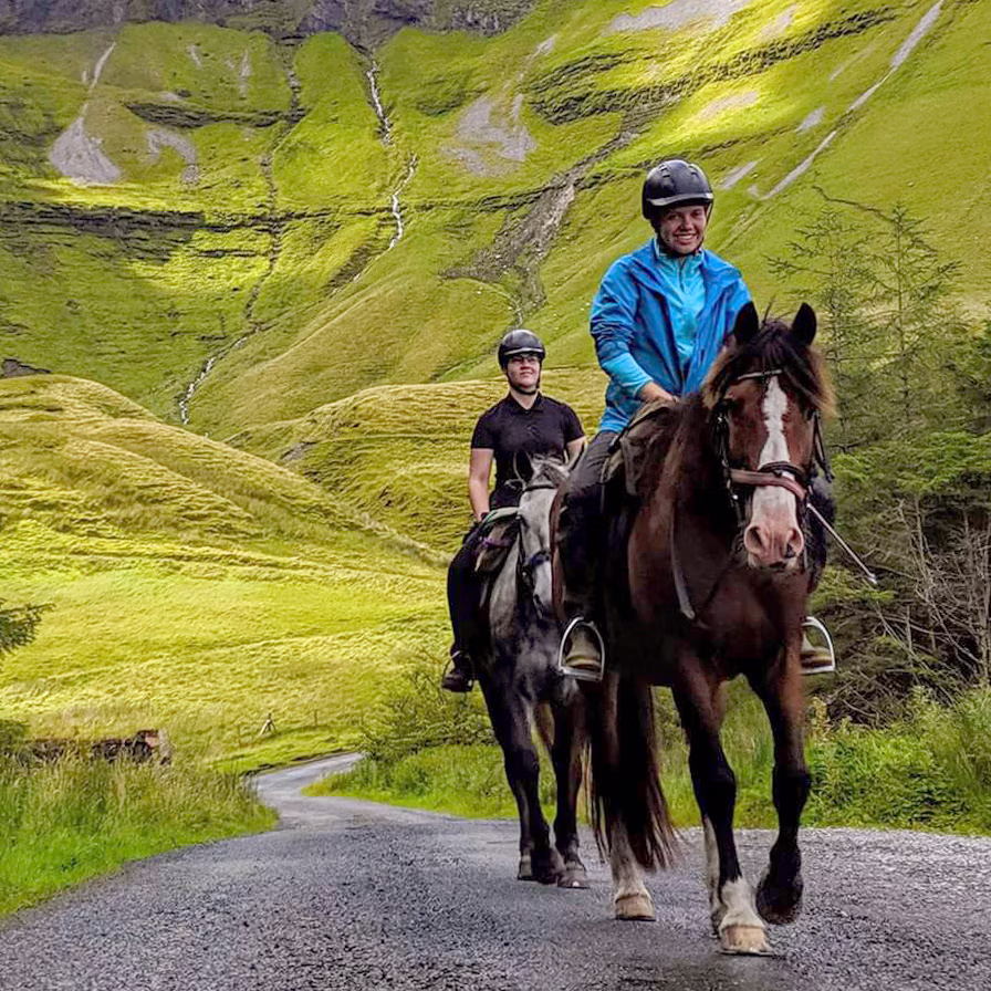 Two horseback riders follow a paved path, framed by a dramatic, vibrant green grass-covered stone wall in the background.