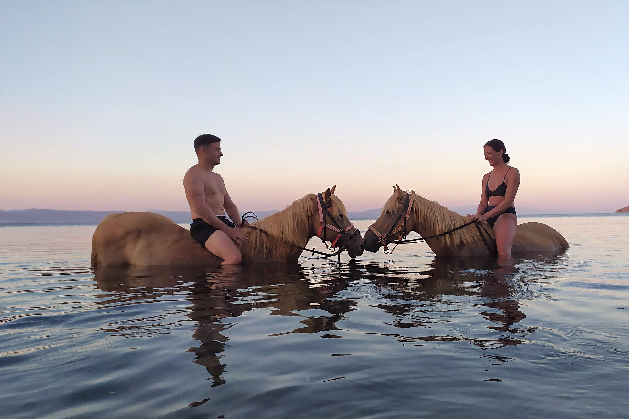 A male and female rider in swimsuits sit on horses facing each other in the water during a warm sunset