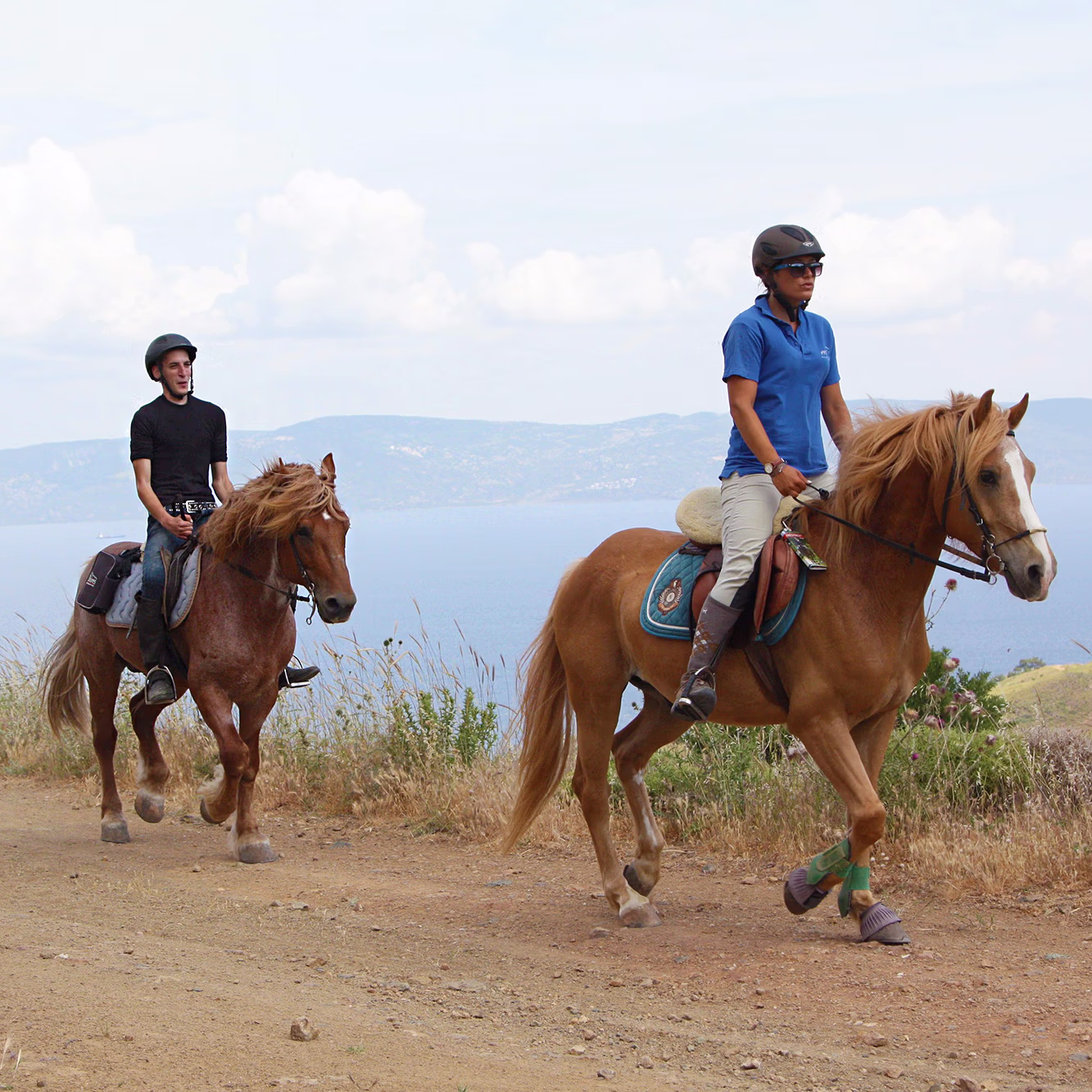 Two horseback riders ascend a scenic hill path with expansive, panoramic views of the ocean in the background.