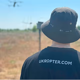 Drone operator in outdoor field wearing branded UKROPTER.COM clothing.

