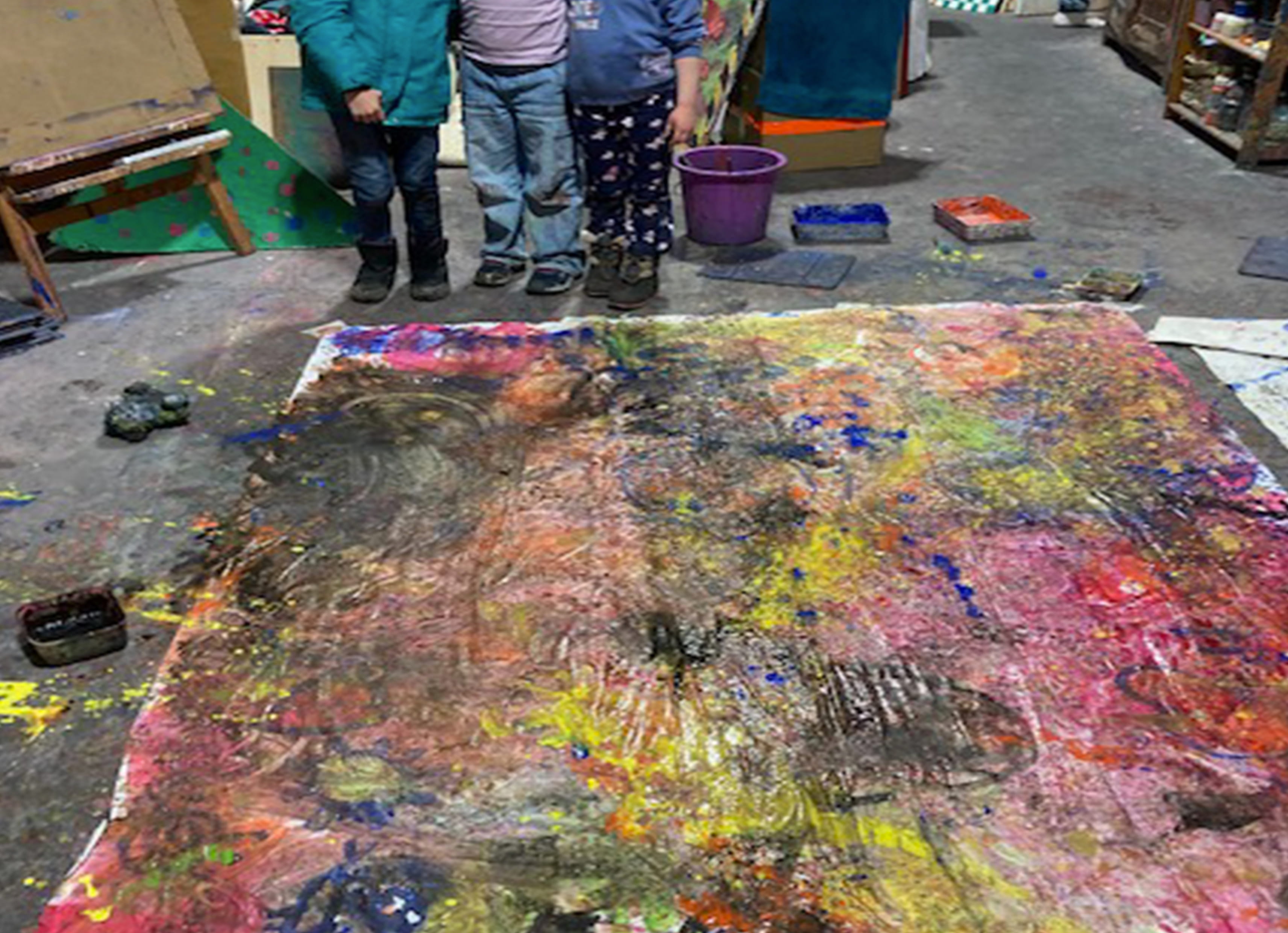 Three children standing behind a large, colorful abstract painting on the floor in an art studio.