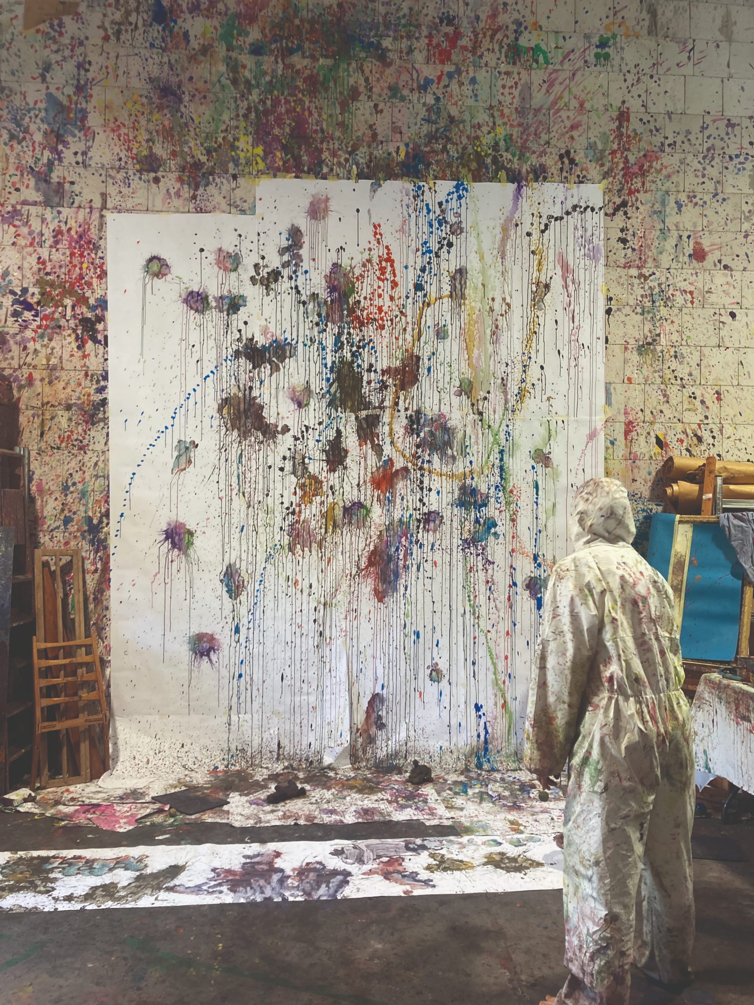 Person in paint-splattered white coveralls standing in front of a large canvas covered with multicolored paint splatters and drips in a cluttered art studio.