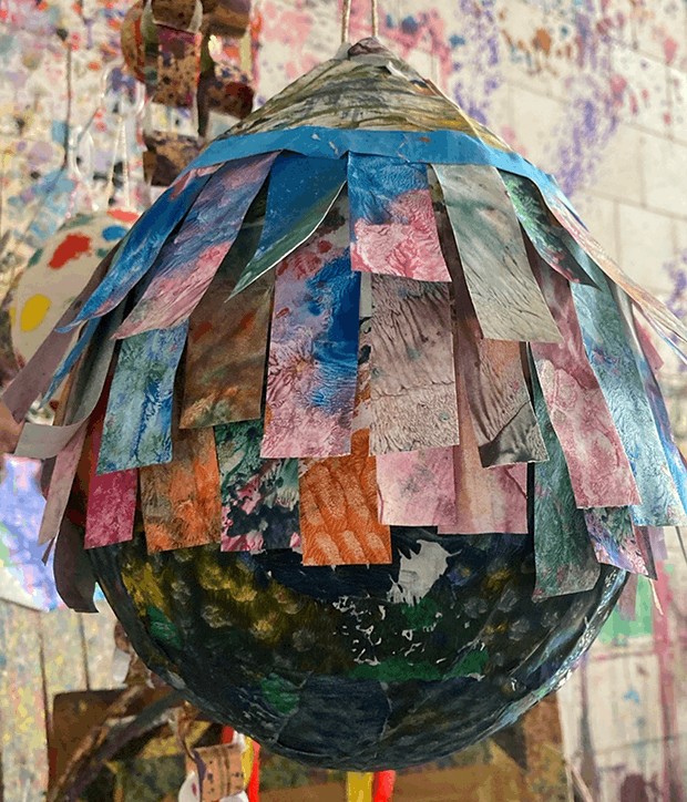 Nahaufnahme einer handgefertigten Piñata aus Pappmaché, verziert mit übereinanderliegenden bunten rechteckigen Papierstreifen in Blau-, Rosa-, Orange- und Grüntönen.