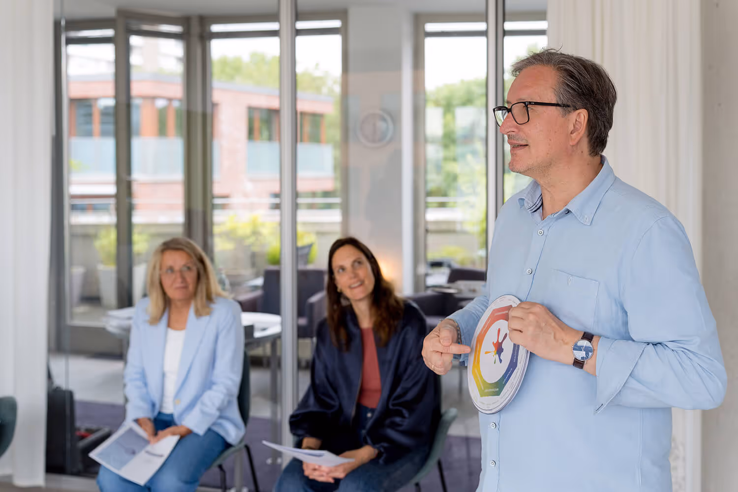 Trainer erläutert ein Modell mit Farbrad; zwei Teilnehmende hören zu (Seminarraum)