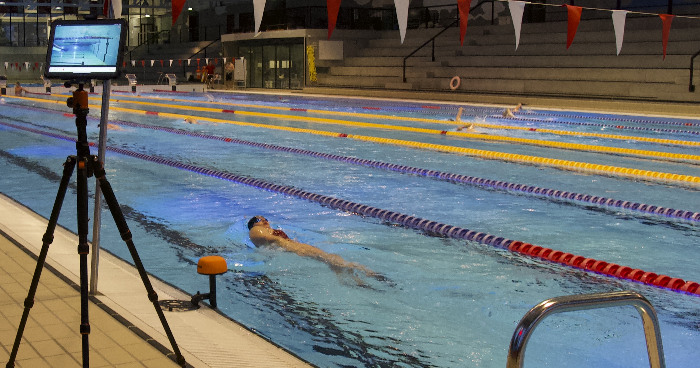 An iPad on a poolside tripod showing live underwater video while a swimmer trains in the lane, with an Athlee camera mounted at the pool edge.