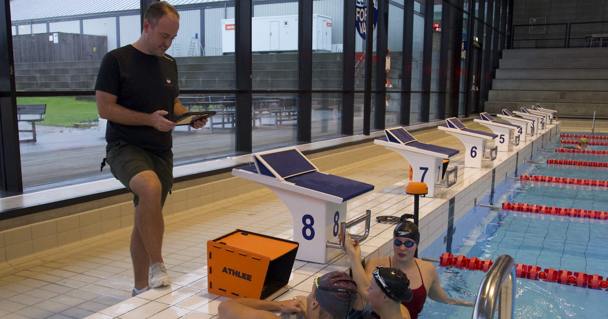 A coach on the pool deck working from an iPad, with the orange Athlee Pool Box at lane 8 and three swimmers at the wall looking up at it.